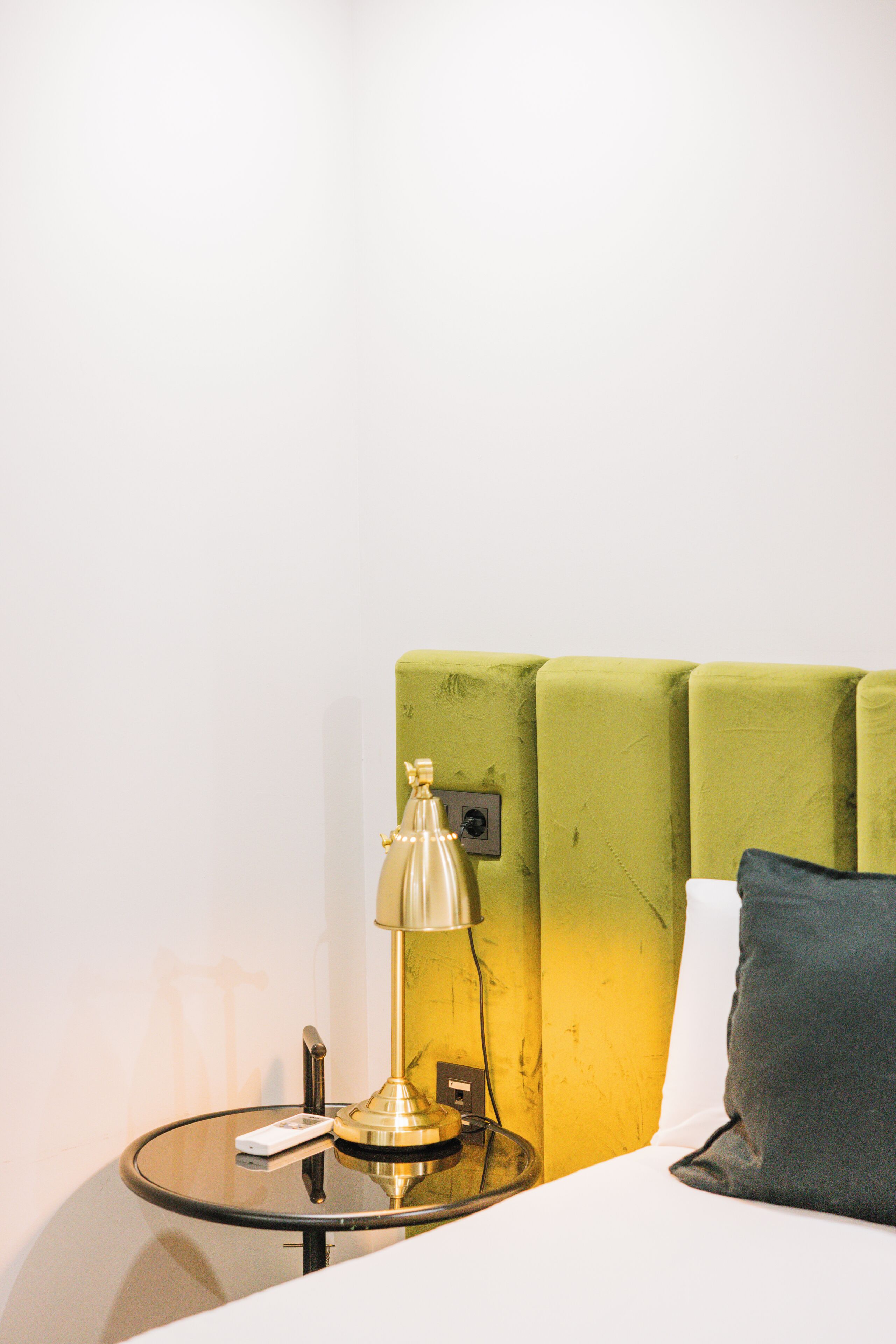Una habitación con un cabecero tapizado en verde, una mesa de vidrio con una lámpara, y una cama blanca con almohada gris.