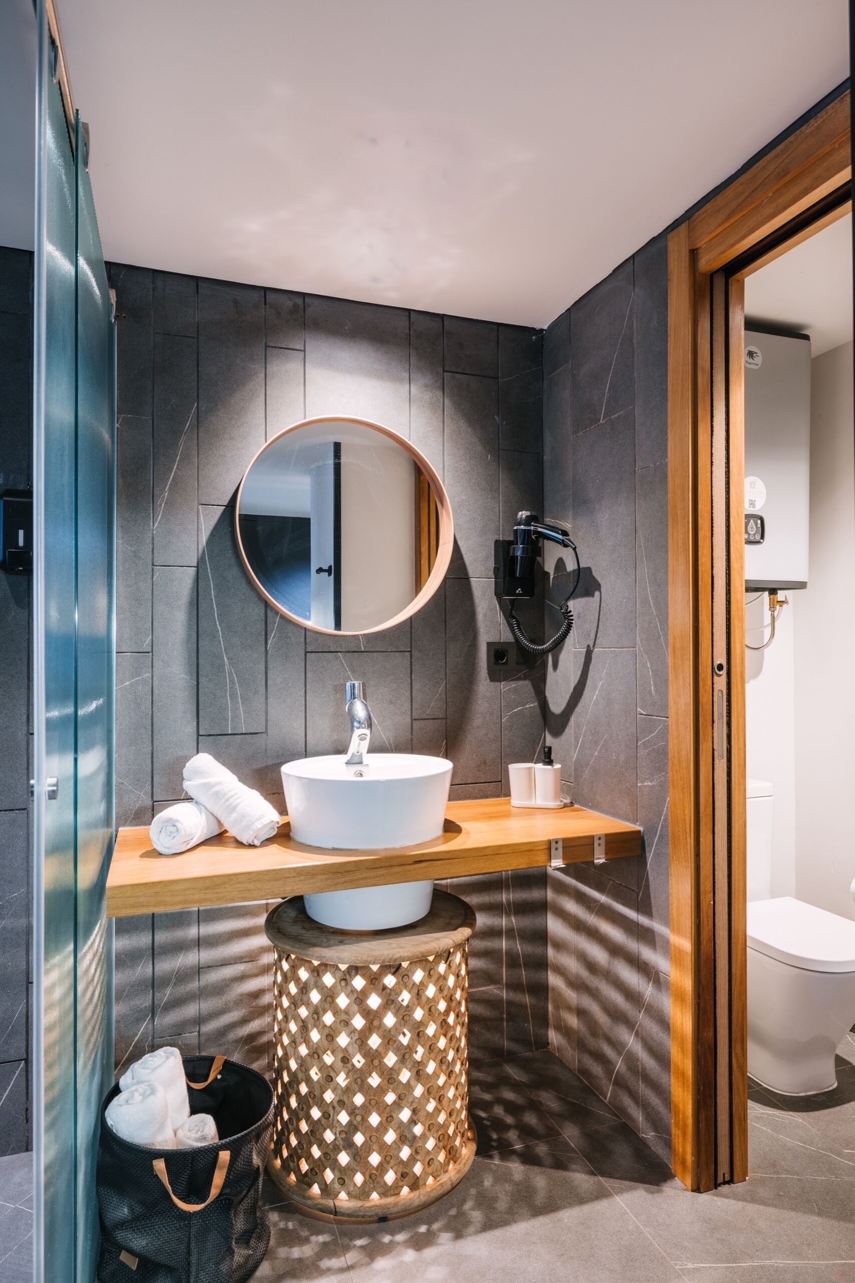Un baño moderno con un espejo redondo, un lavamanos blanco, una toalla, un cubo de basura y una zona de ducha.