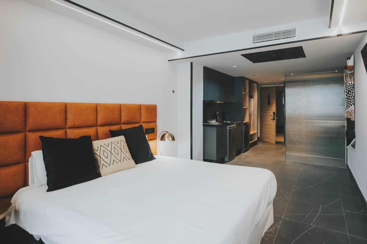 Ein modernes Hotelzimmer mit einem großen Bett, einem Kopfteil, einem Nachttisch, einer Lampe und Blick in den Küchenbereich.