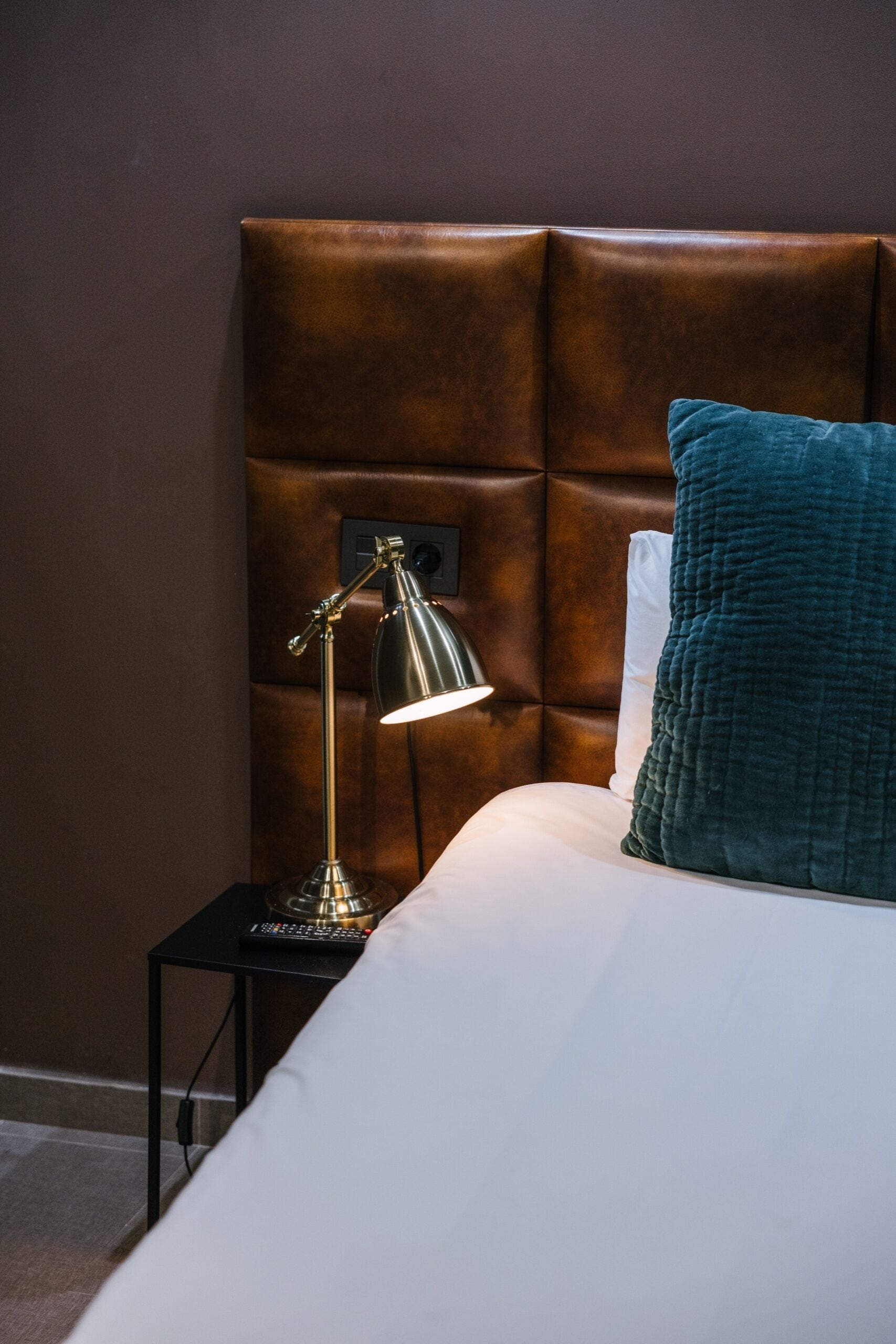 Una cama con cabecera, una lámpara de noche, un control remoto y una almohada color turquesa.