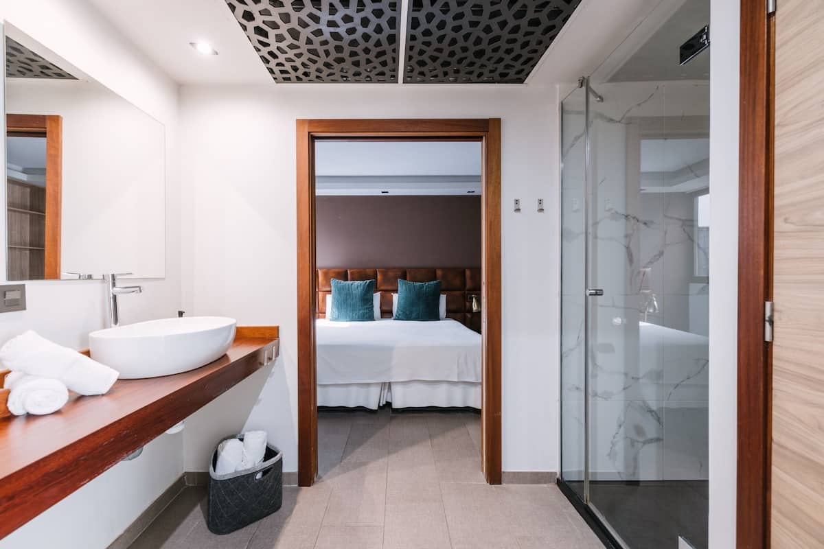 Ein modernes Hotelzimmer mit einem großen Bett, einem Badezimmer mit Glasdusche und einem Holzanrichte mit Waschbecken.