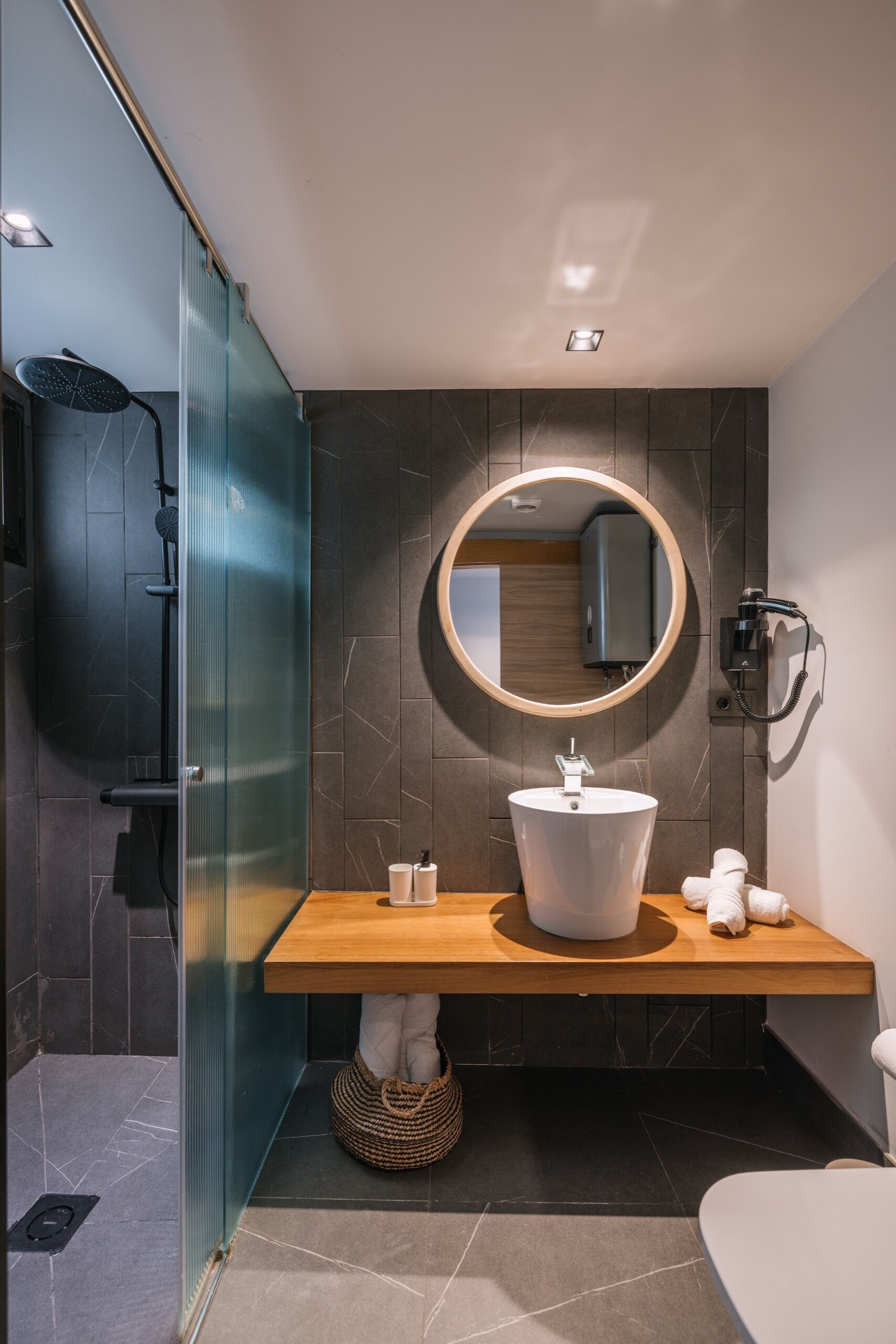 A modern bathroom with a glass shower enclosure, a wooden vanity, a round mirror, and a toilet.