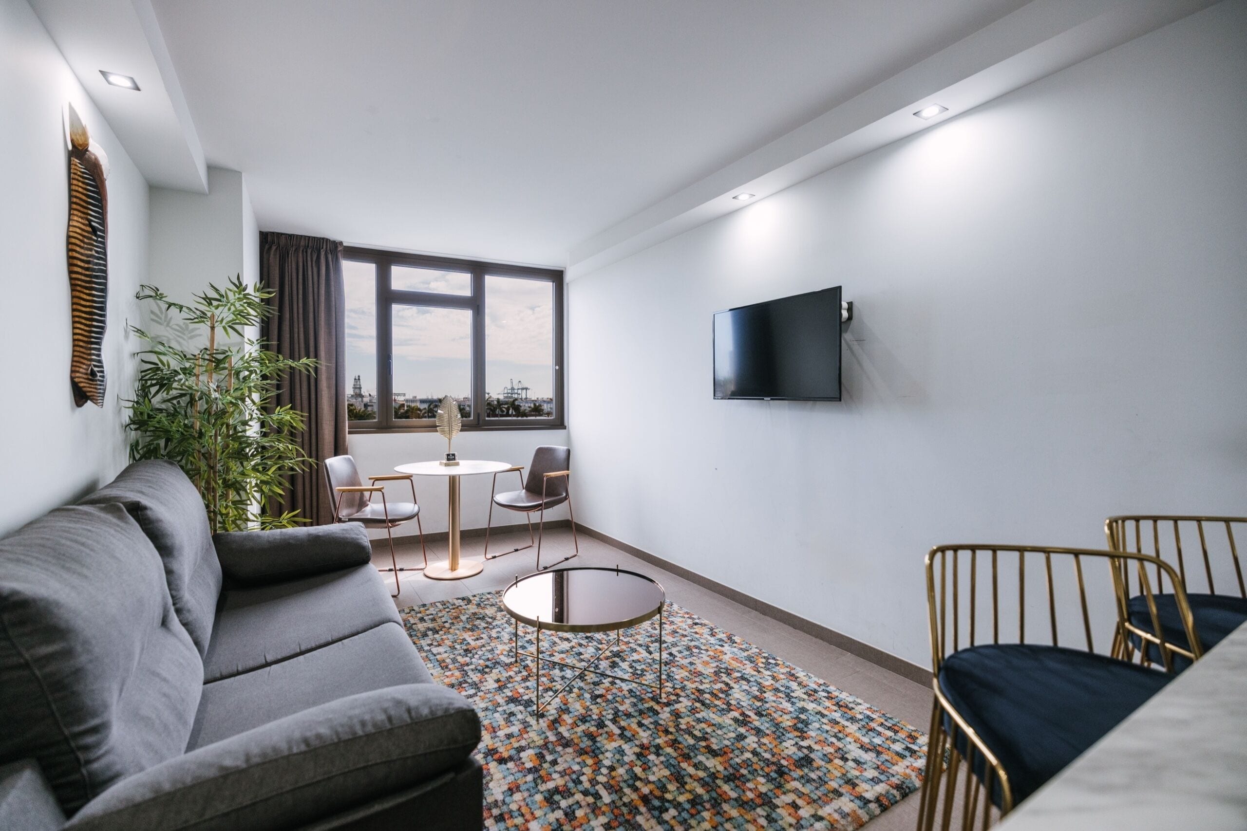 A modern living room with a sofa, a round coffee table, a TV mounted on the wall, and a large window with a city view.