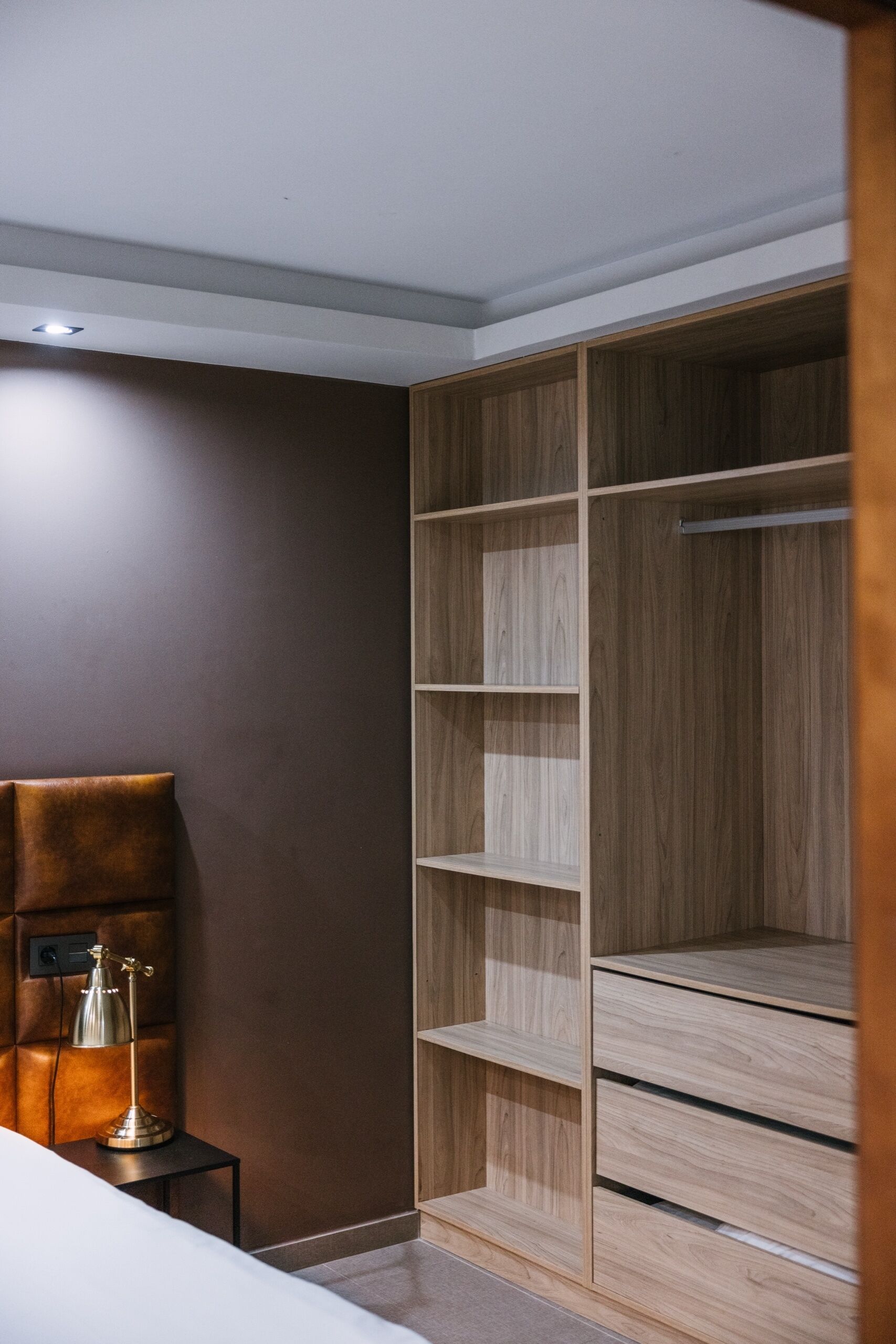 A bedroom with a bed, a nightstand, a lamp, a wooden wardrobe, and a brown wall.