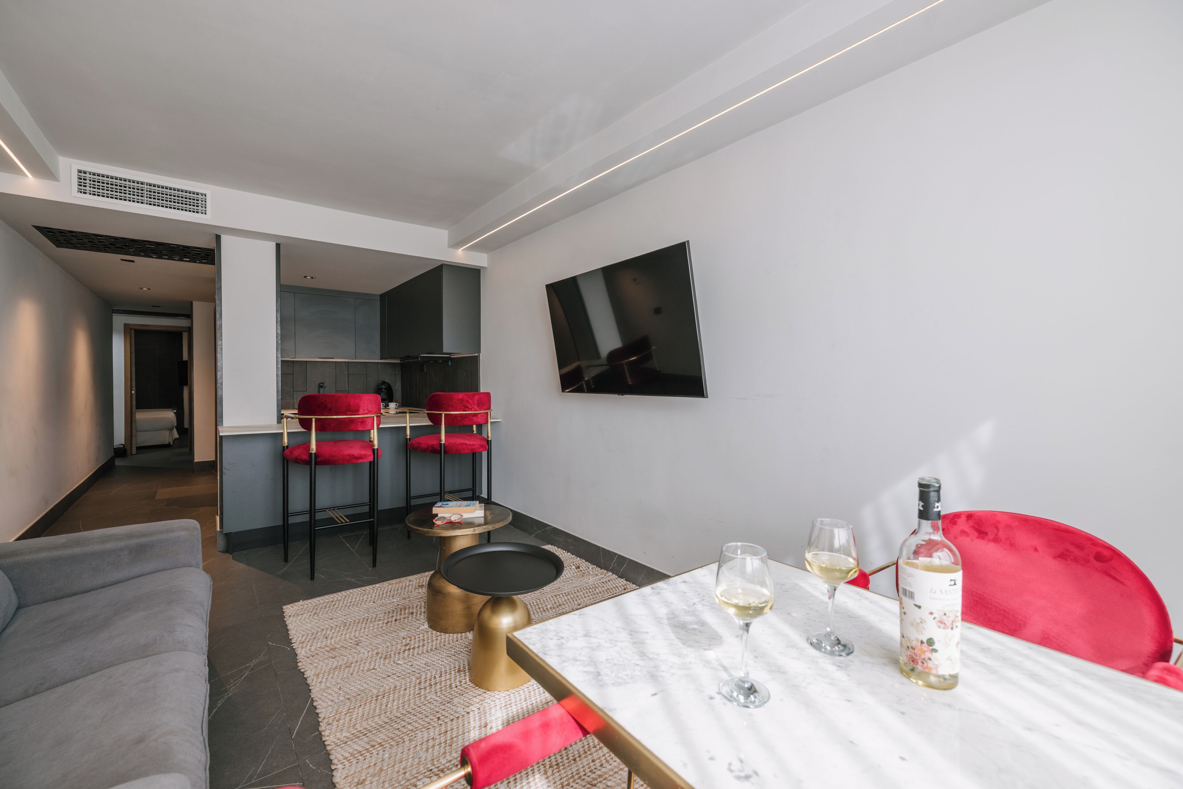 A modern living room with a sofa, a dining table with chairs, a kitchen area, and a TV mounted on the wall.
