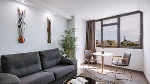 Een moderne woonkamer met een grijze bank, een rond salontafel, een kleine ronde tafel met twee stoelen, een groot raam met uitzicht, en twee dierenhoofds aan de muur bevestigd.