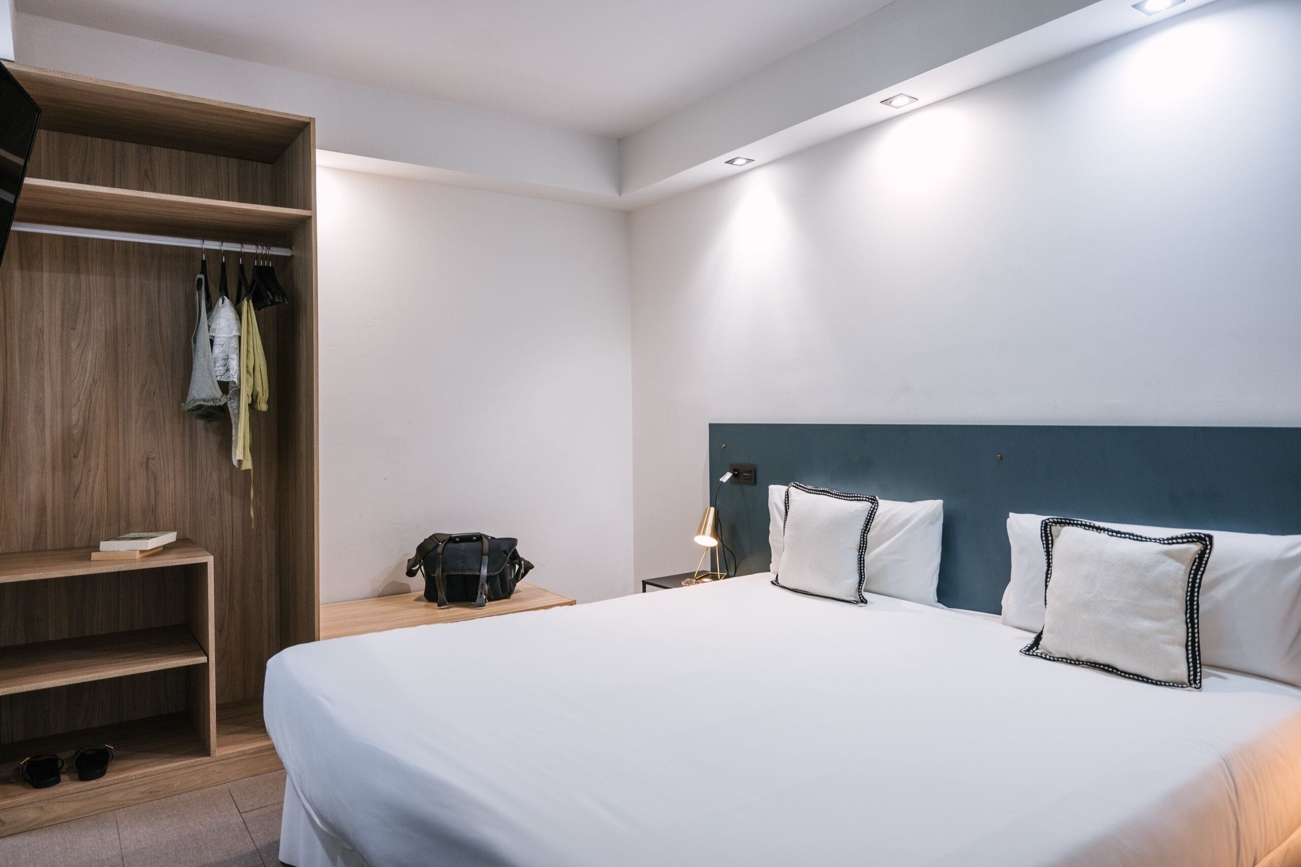 A hotel room with a bed, a wooden wardrobe, a bedside table with a lamp, and a nightstand.