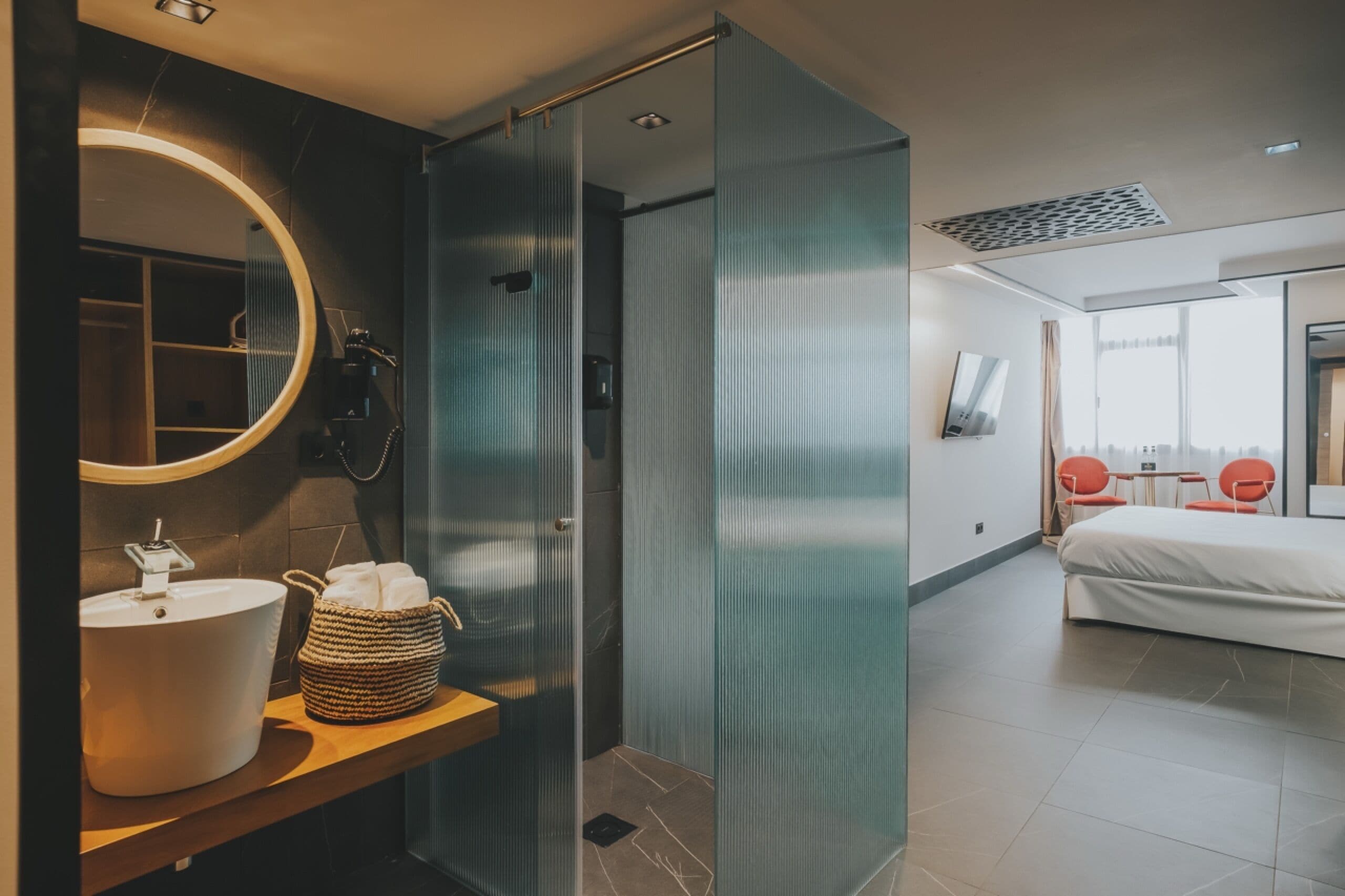 A modern hotel room with a large round mirror, a sink, a shower, a bed, and a small table with chairs.