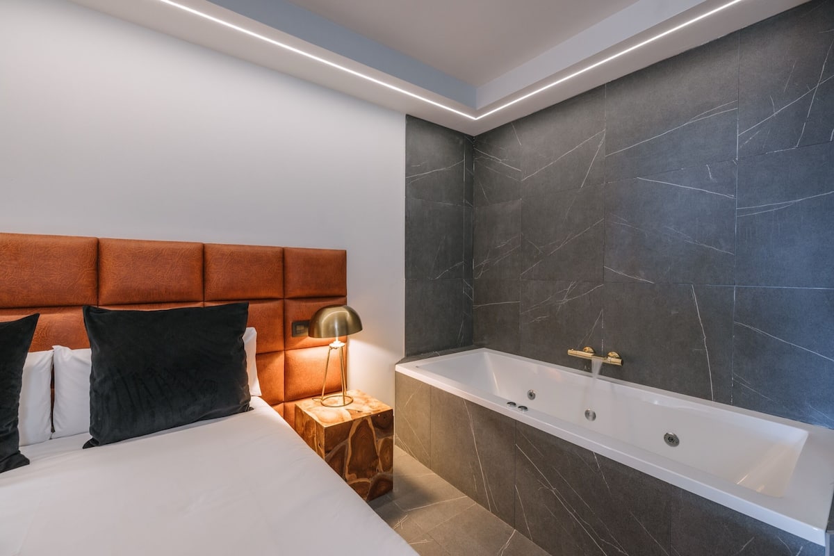 Ein modernes Badezimmer mit einer Badewanne, einem Holfnachtkästchen mit einer Lampe und einem Kopfteil mit Kissen.