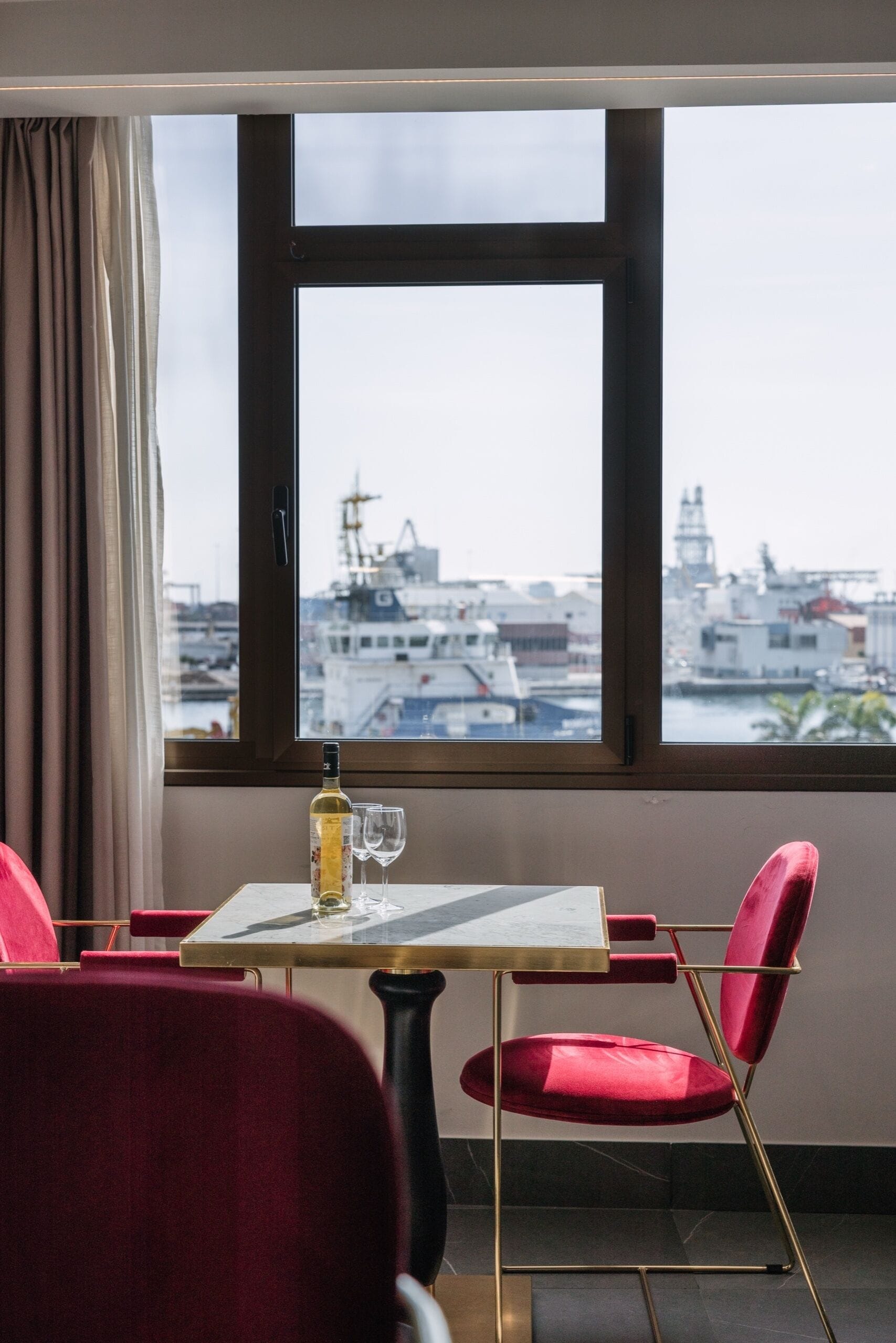 A room with a table and chairs, a bottle and glass on the table, and a view of a marina through large windows.
