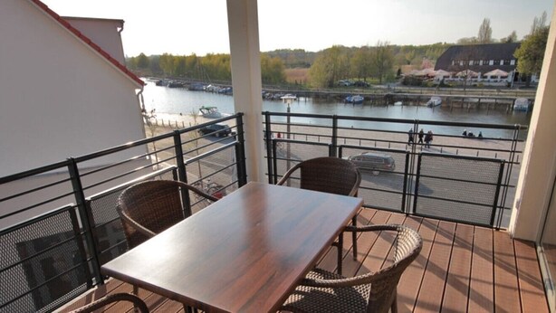 A balcony with a table and chairs, overlooking a river with boats and a building in the background. - Modern furnished apartment (Greifswald)