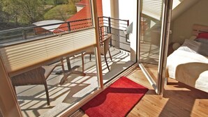 A bedroom with a bed, a red rug, a balcony with a chair, and a view of a red-roofed building. - Modern furnished apartment for the family (Greifswald)