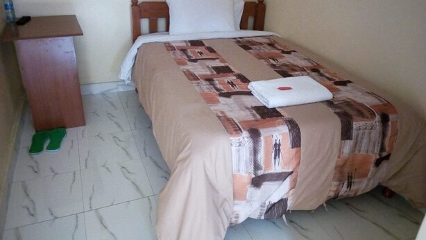 A single bed with a patterned bedspread, a wooden headboard, a bedside table, and a pair of green slippers on the floor. - Sweet Hut Hotel (Marua)