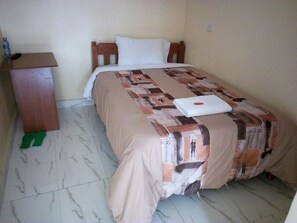 A single bed with a patterned bedspread, a wooden headboard, a bedside table, and a pair of green slippers on the floor. - Sweet Hut Hotel (Marua)