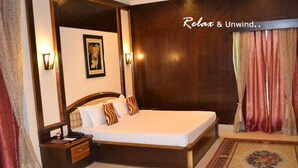 A hotel room with a large bed, wooden headboard, two bedside tables, a nightstand, a framed picture on the wall, and a window with curtains. - Hotel Rudra Continental Rudrapur (Kichha)