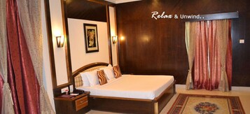 A hotel room with a large bed, wooden headboard, two bedside tables, a nightstand, a framed picture on the wall, and a window with curtains.