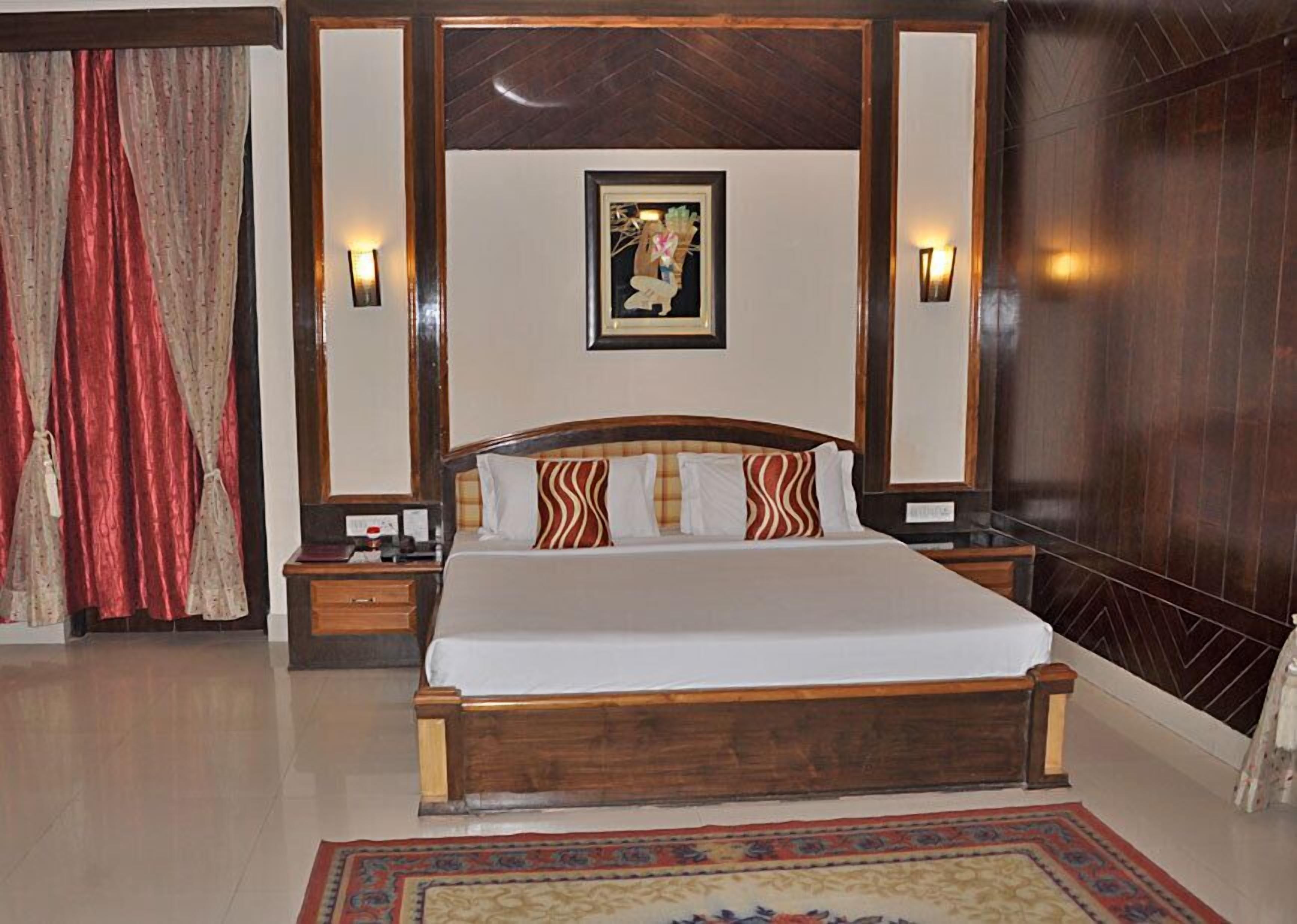 A hotel room with a large bed, wooden headboard, two bedside tables, a nightstand, a wall-mounted picture, and a patterned rug.