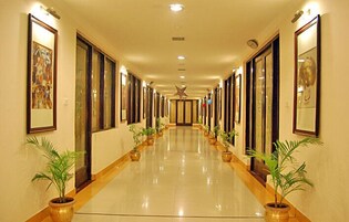A long, well-lit hallway with framed artwork on the walls, potted plants along the sides, and a shiny floor.
