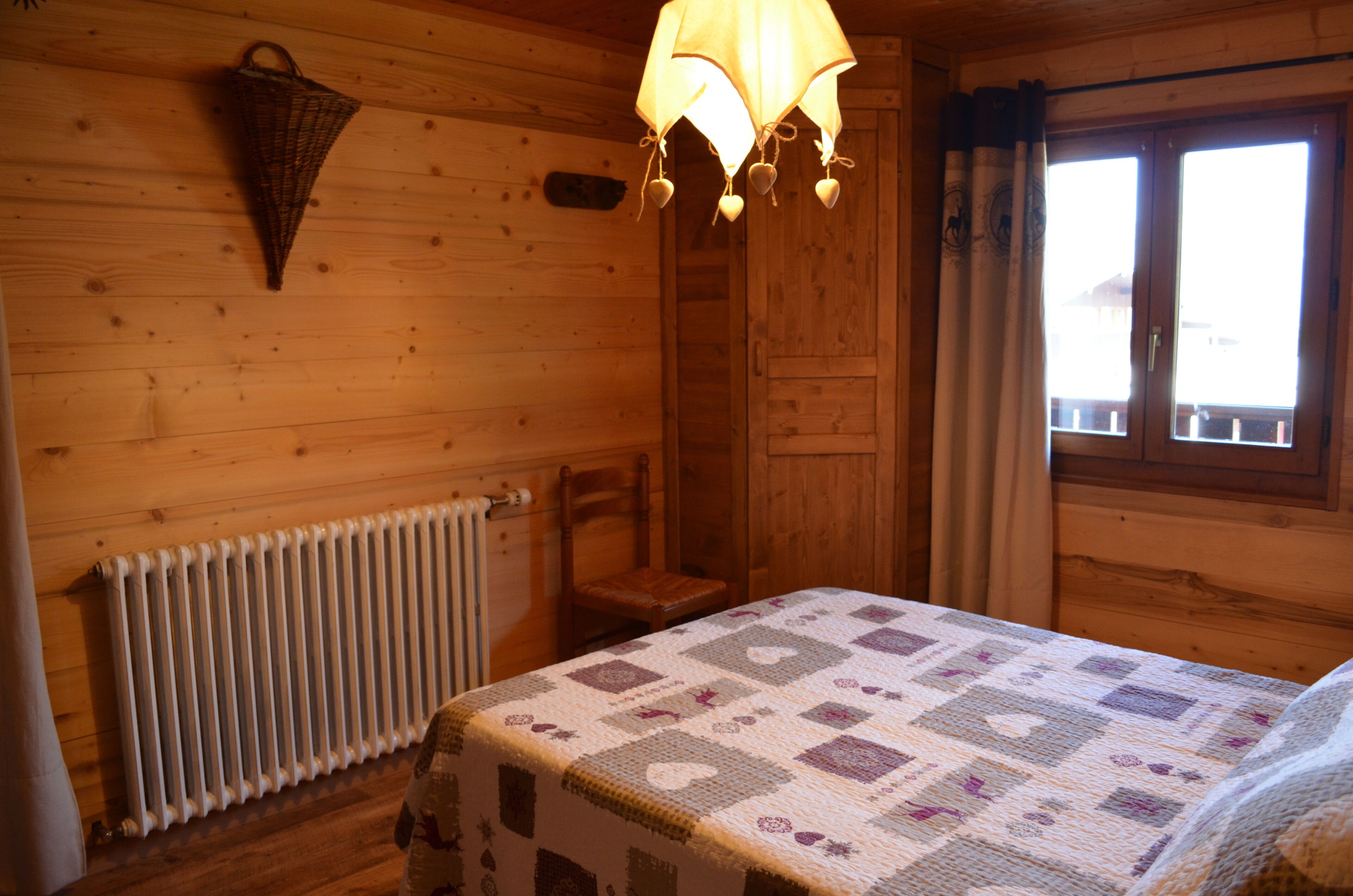 A wooden cabin room with a bed, a chair, a hanging light fixture, a radiator, and a window with curtains.