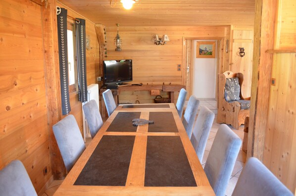 A wooden interior with a long dining table, chairs, a television, and a small kitchen area.
