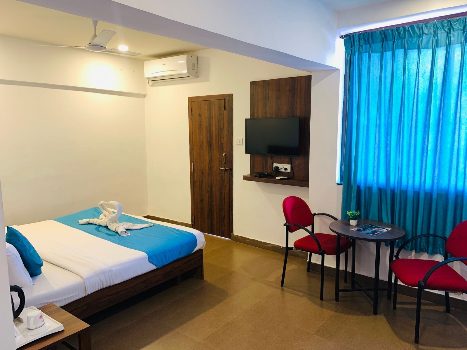 A hotel room with a bed, a desk with a chair, a TV mounted on the wall, and a window with curtains.