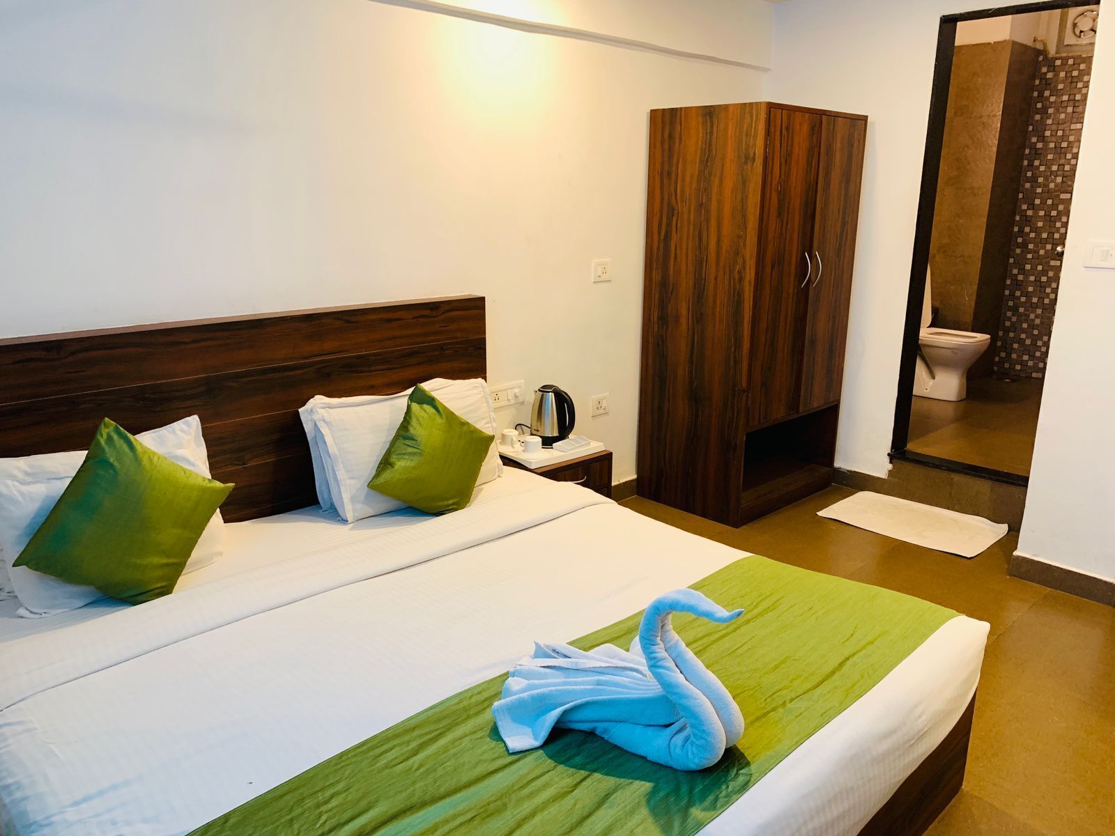 Ein Hotelzimmer mit einem Bett, grünen und weißen Kissen, einem Holzschrank, einem Nachttisch mit einem Wasserkocher und einem zu einem Schwan geformten Handtuch.
