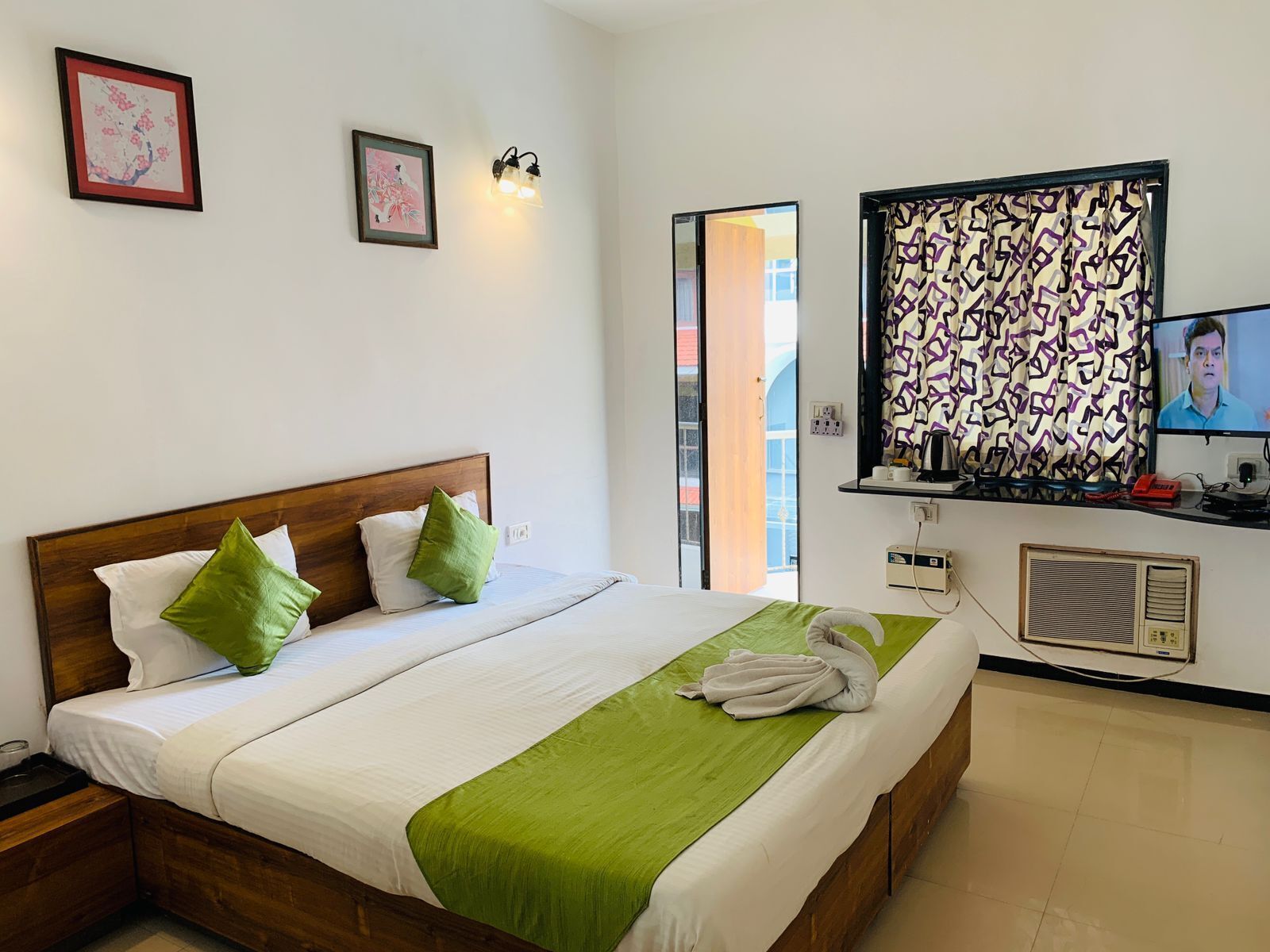 Een hotelkamer met een groot bed, twee kussens, een groene plaid, een televisie en een raam met patroon gordijnen.