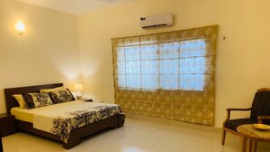 Une chambre d’hôtel comprenant un lit, une chaise, une table, une fenêtre avec un rideau, un climatiseur et un luminaire fixé au mur.