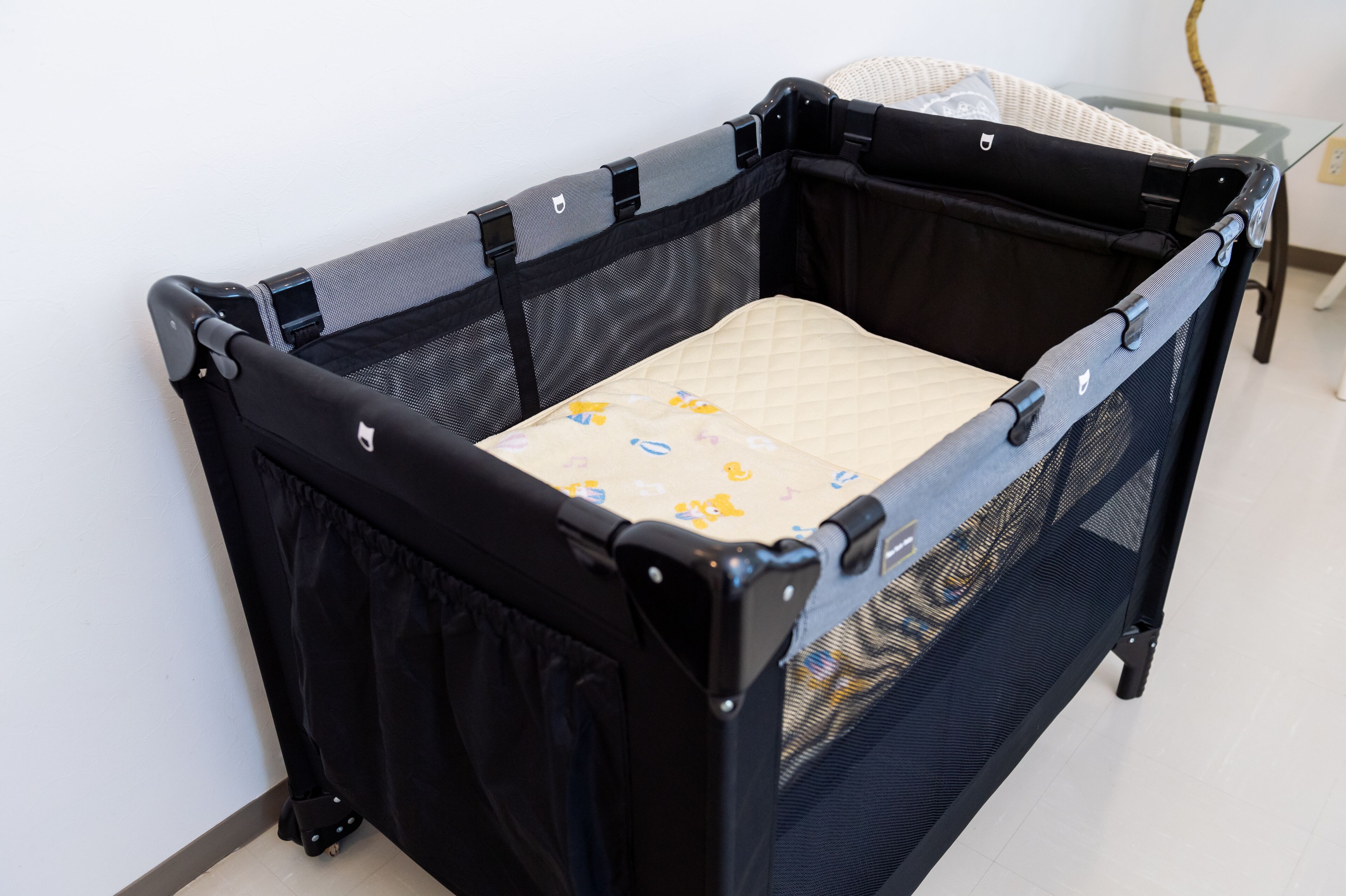 A portable crib with a mesh side and a quilted mattress, placed against a white wall.