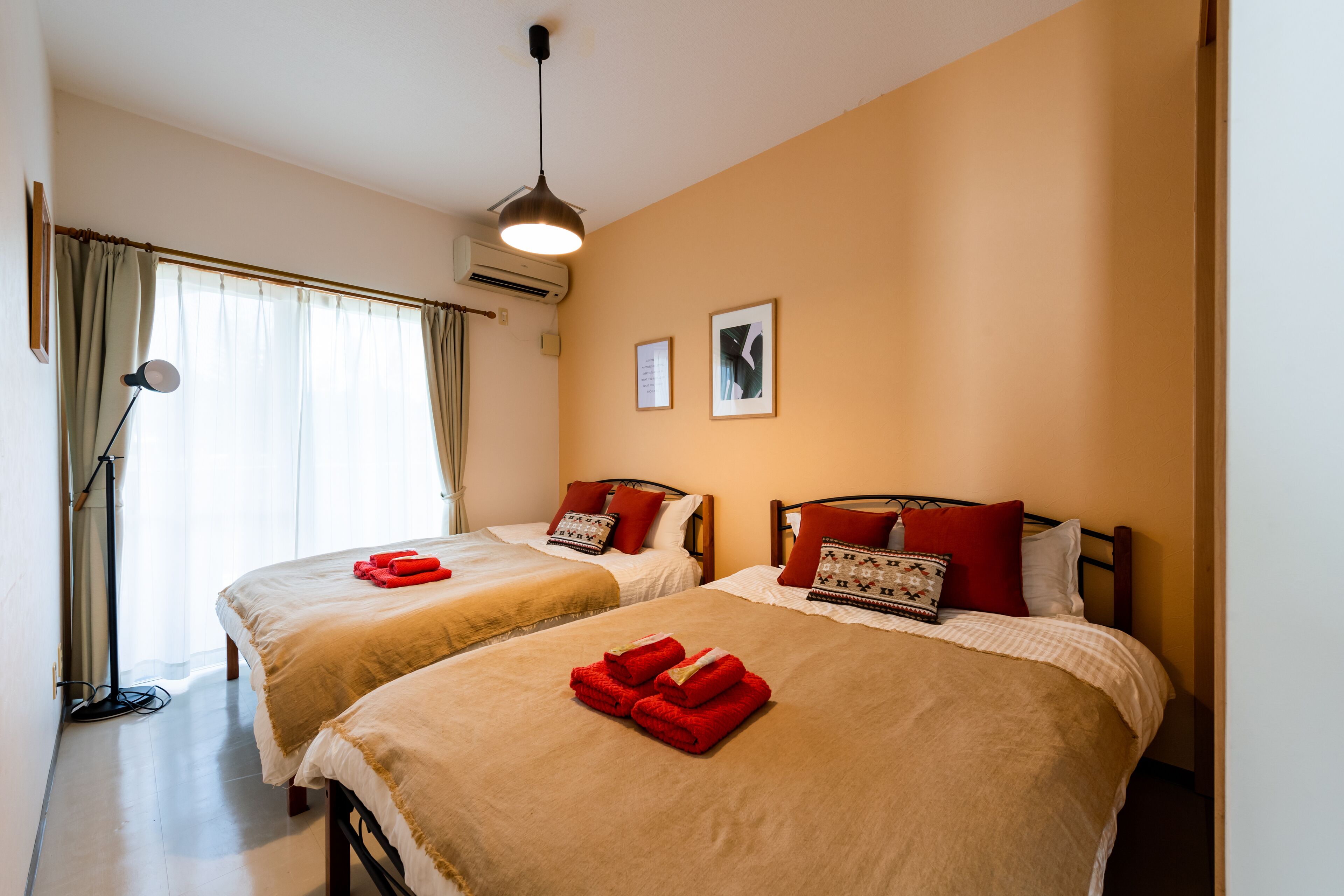 A hotel room with two beds, each with red towels, a standing lamp, and framed pictures on the wall.