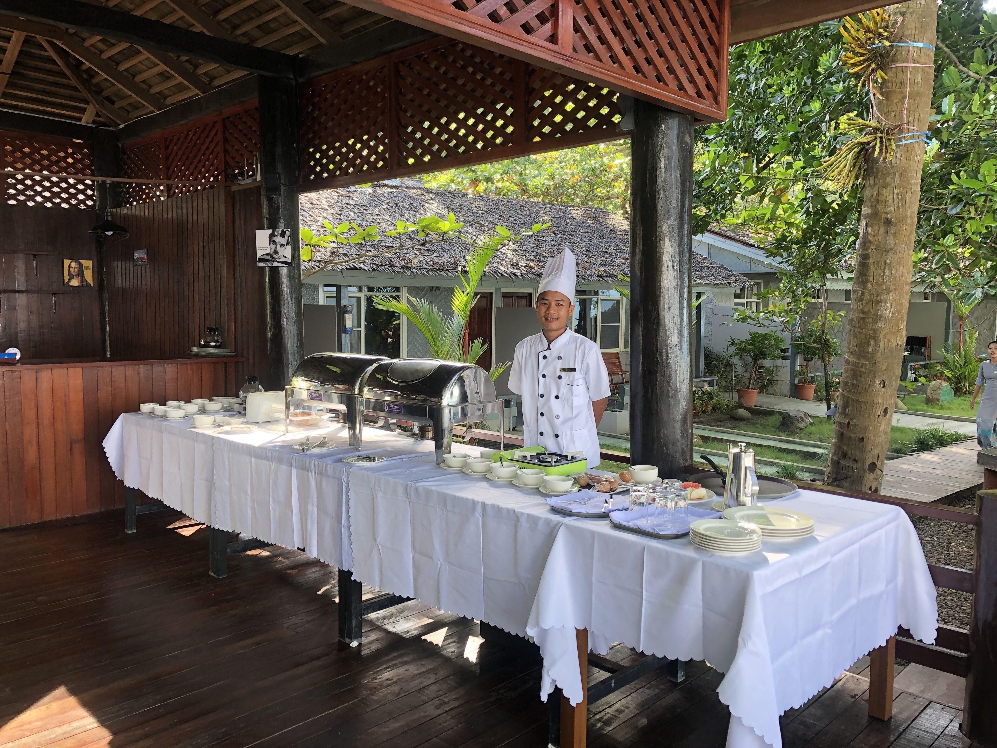 A covered outdoor dining area with a buffet setup, including a chef, a wooden structure with lattice panels, and a view of a house and greenery in the background.