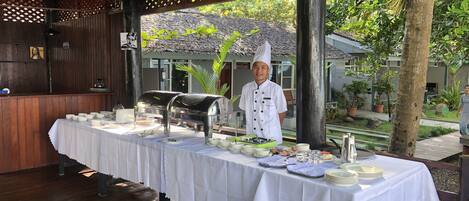 Un área de comedor al aire libre techada, con mesas de buffet, un chef, una estructura de madera con paneles de celosía y vistas a una casa y follaje al fondo.