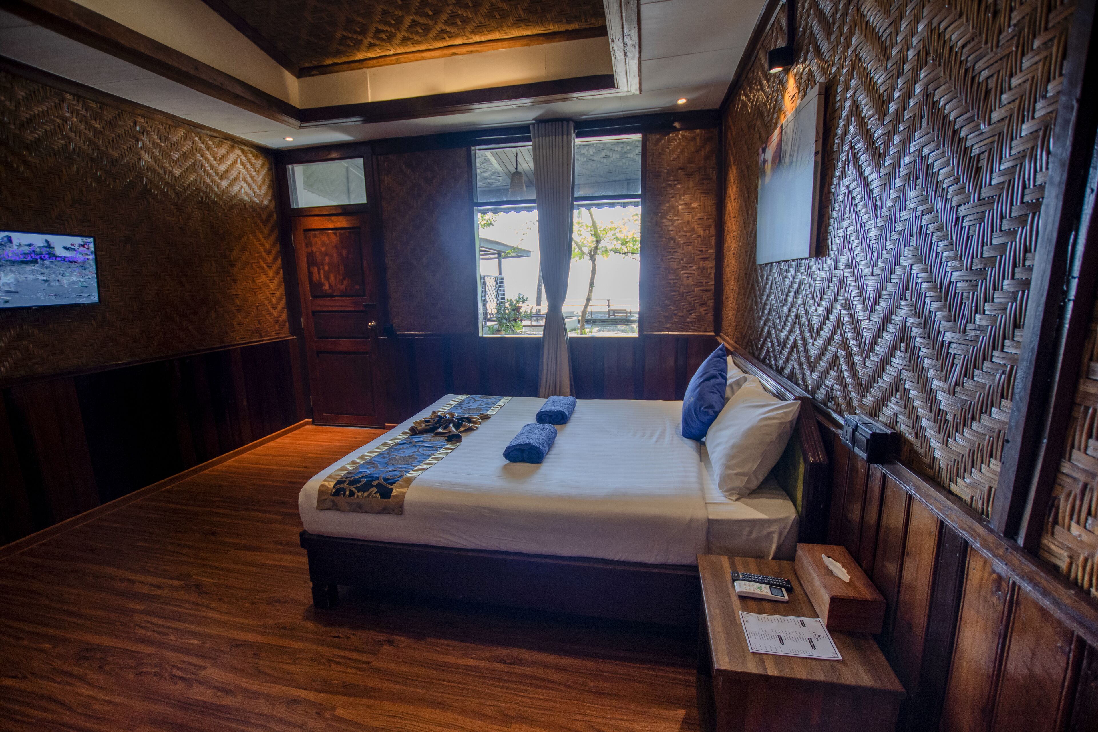 Habitación de hotel con piso de madera, una cama con tablero de cabecera estampado, una mesita de noche con lámpara y un ventanal con vista al exterior.