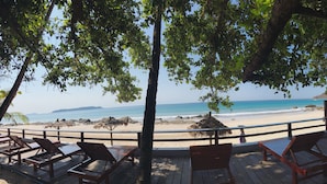 Eine Holzterrasse mit Liegestühlen mit Blick auf einen Strand, an dem sich eine Reetdachhütte und eine felsige Küste befinden.
