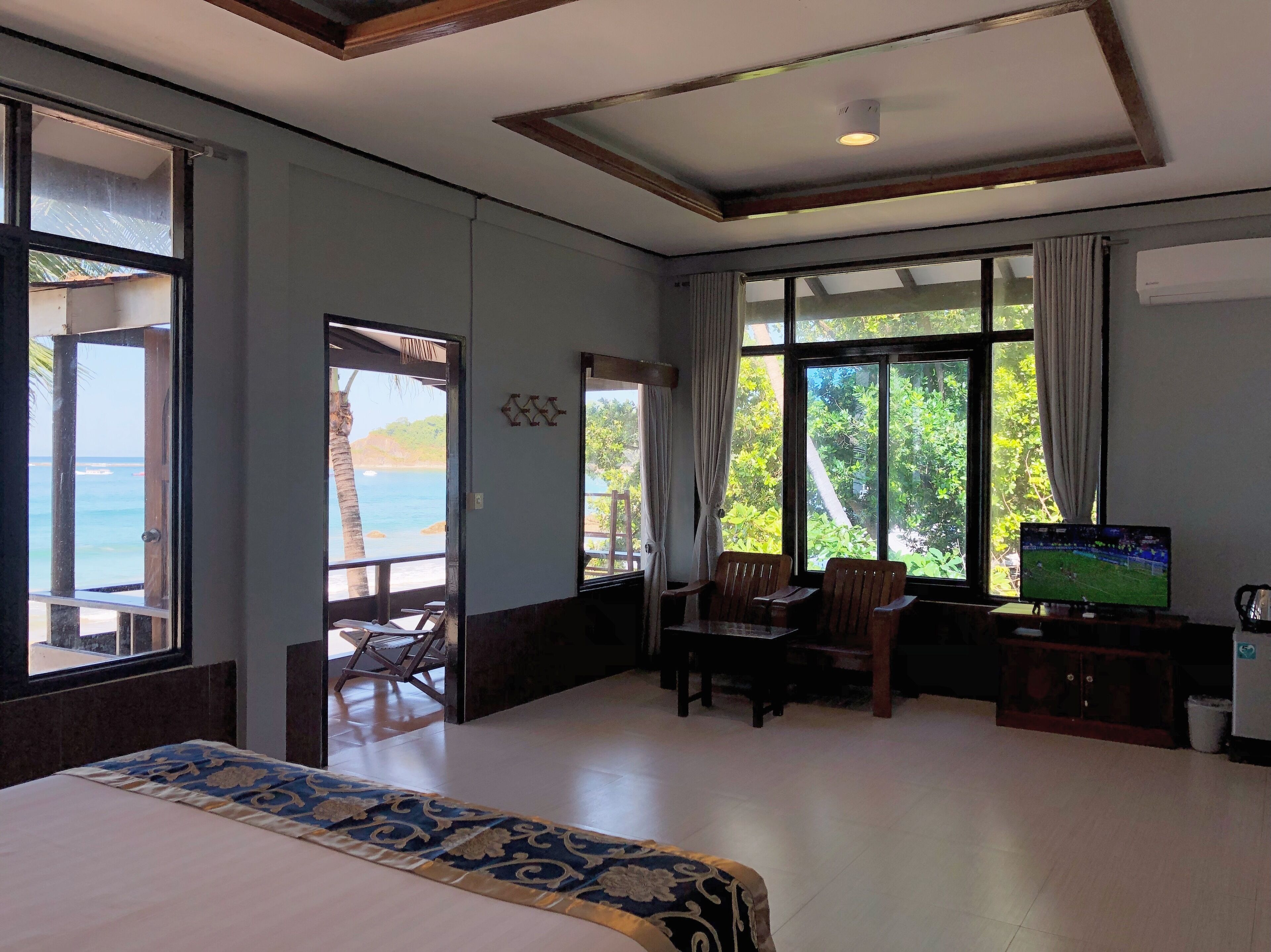 A hotel room with a bed, a desk with a TV, and large windows offering a view of the sea.