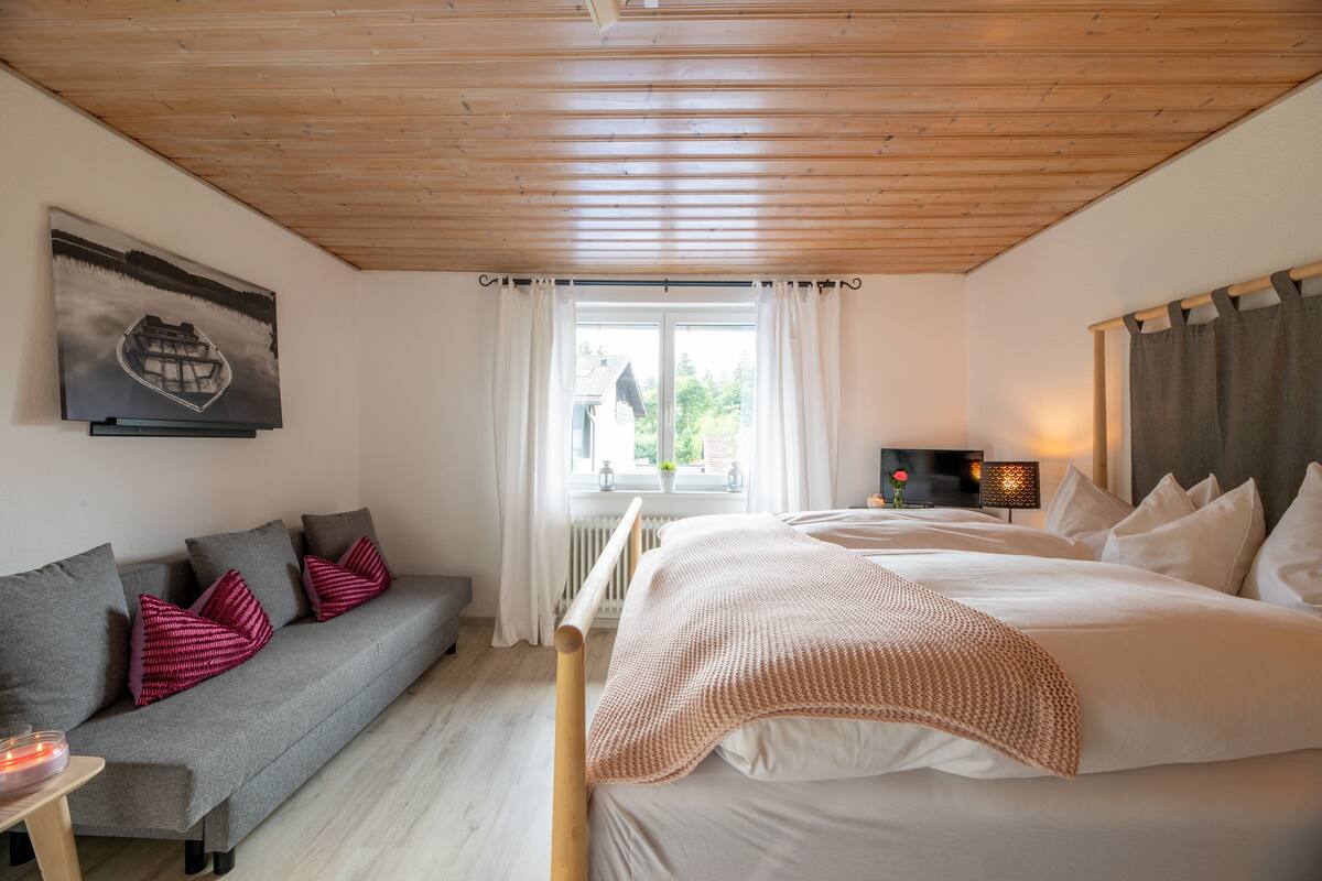 Quarto com teto de madeira, sofá cinza com almofadas vermelhas, cama com roupa de cama branca, janela com cortinas e um quadro na parede.