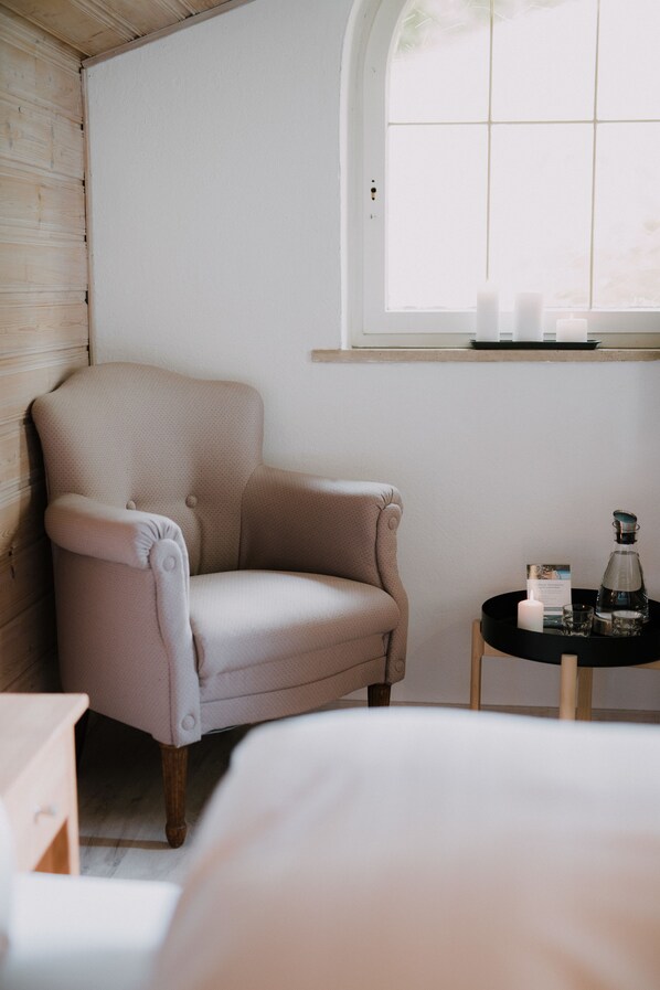 A room with a beige armchair, a small round table with a candle and a bottle, and a window with a view.