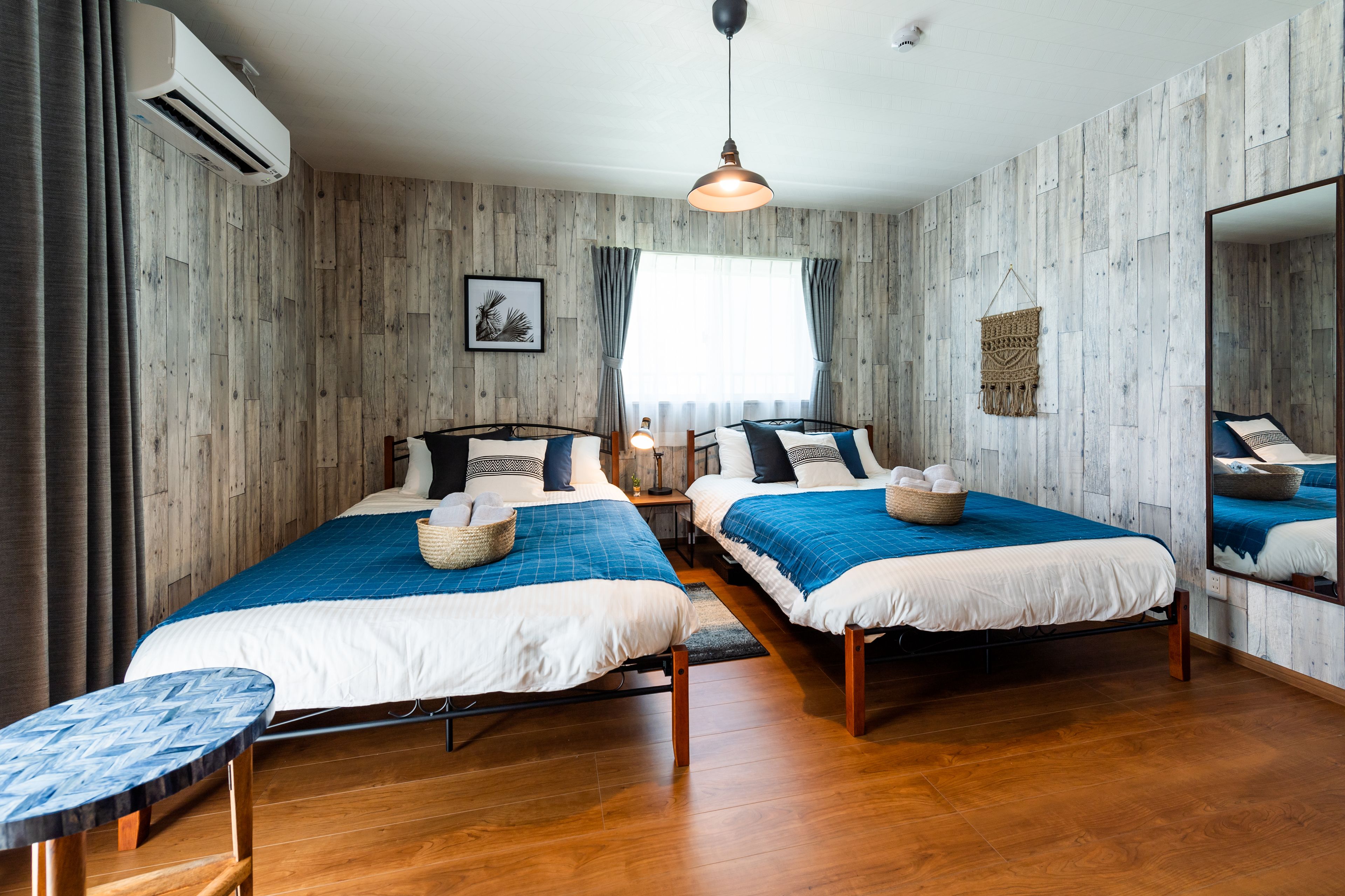 A room with two beds, wooden flooring, a hanging light fixture, and a large mirror.
