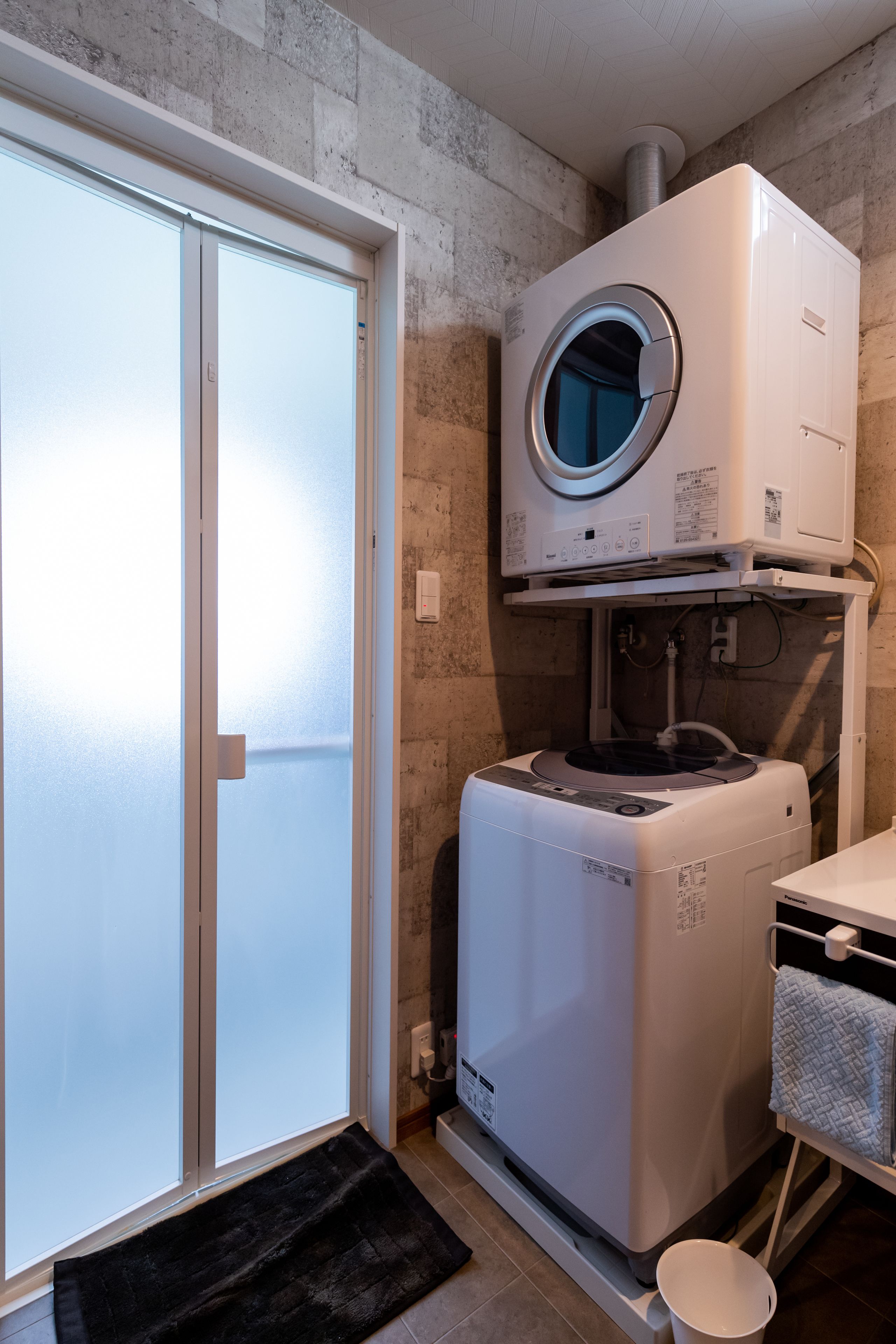 A compact laundry room with a front-loading washer and dryer, a towel rack, and a door leading to a balcony.