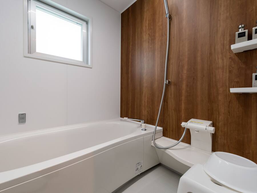 A modern bathroom with a white bathtub, a shower, and a toilet. There is a window above the bathtub and wooden paneling on the wall.