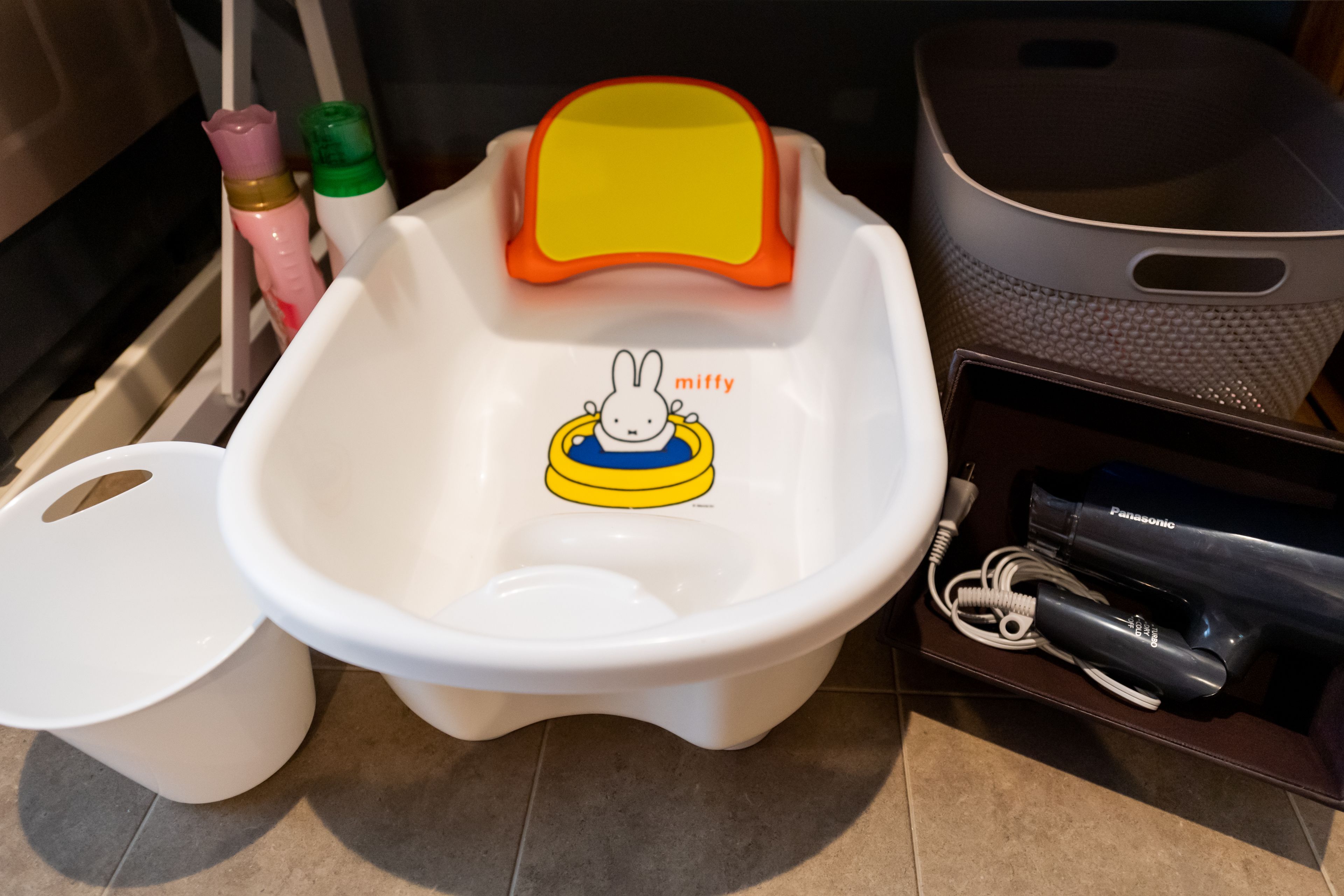 A white baby bathtub with a cartoon character and the word "miffy" on it, placed on a tiled floor next to a white trash bin and a black Panasonic hairdryer.
