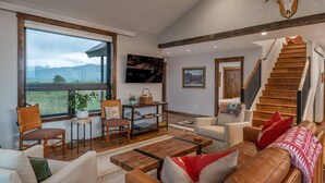 Ein geräumiges Wohnzimmer mit großem Fenster, einer Holztreppe und einem aufgehängten Damhirschschädel an der Wand.