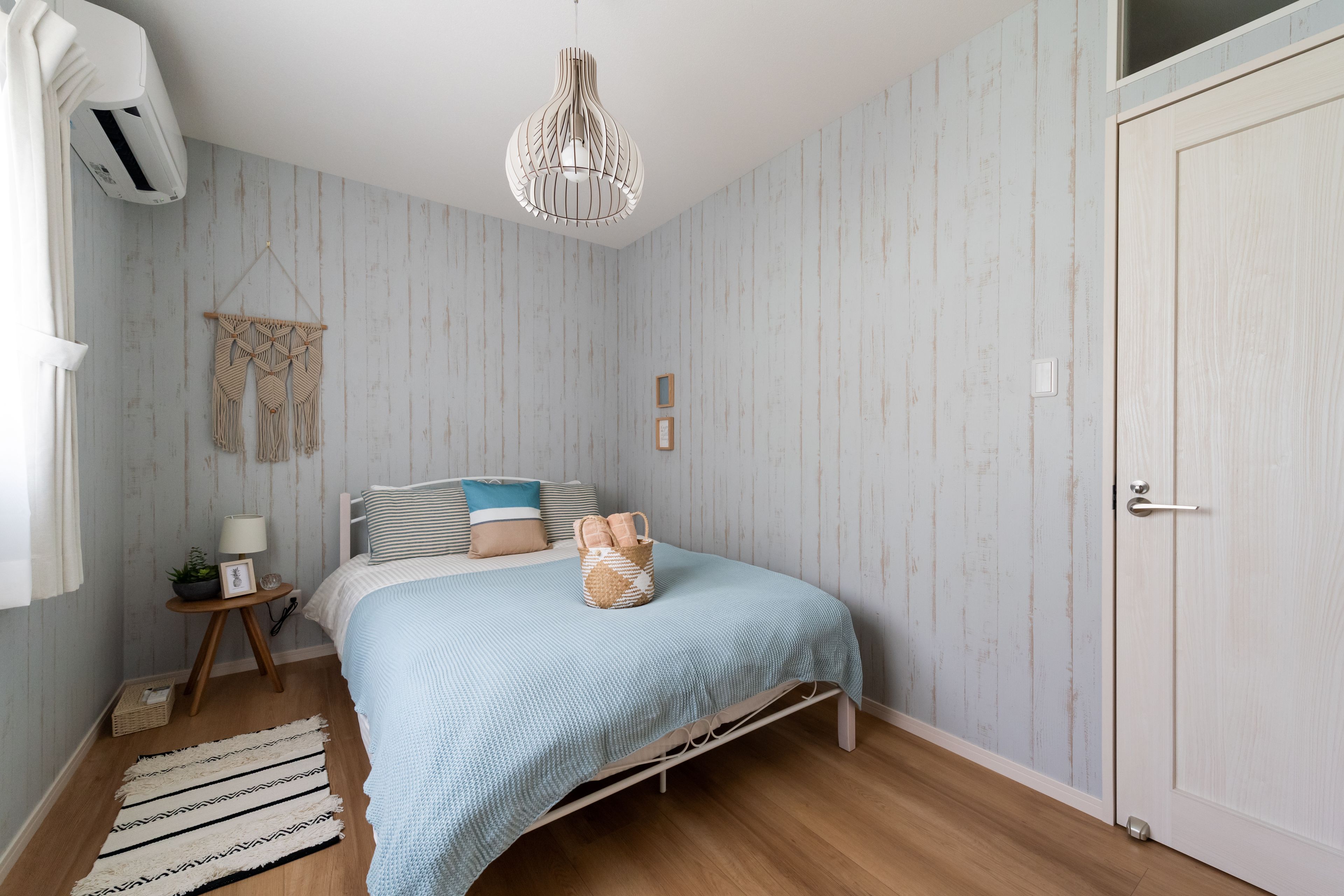 Un dormitorio con una cama, un adorno de macramé en la pared, una mesita de noche con lámpara y suelo de madera.