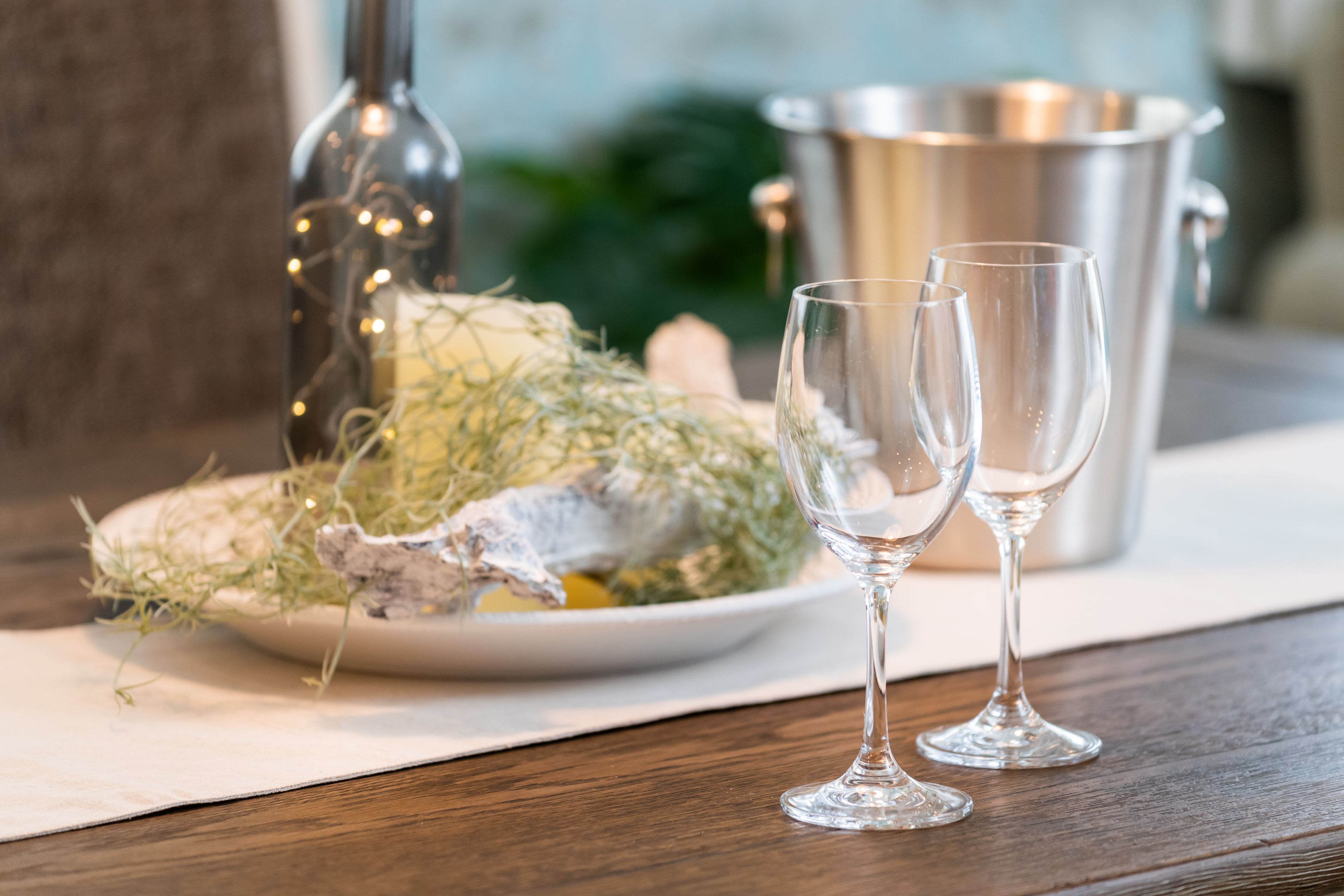A wooden table with a plate of food, two empty wine glasses, a bottle, and a metal container.