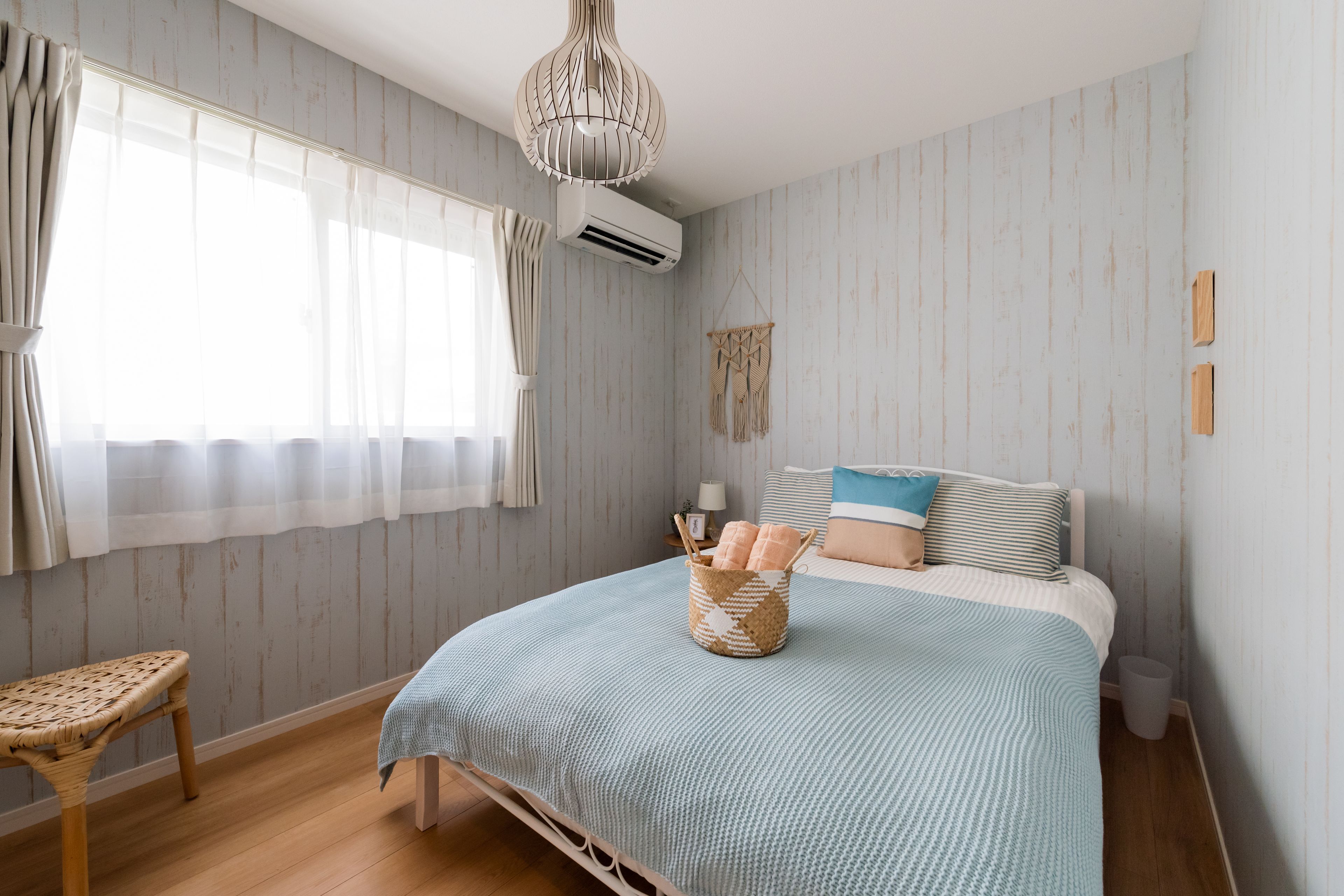 A bedroom with a bed, a wicker chair, a hanging macrame wall decoration, and an air conditioning unit.