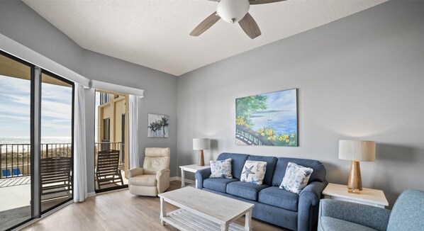 A modern living room with a blue sofa, a beige armchair, and a white coffee table. There is a painting of a landscape on the wall, and a ceiling fan above. - Phoenix V 110 by Liquid Life (Orange Beach)