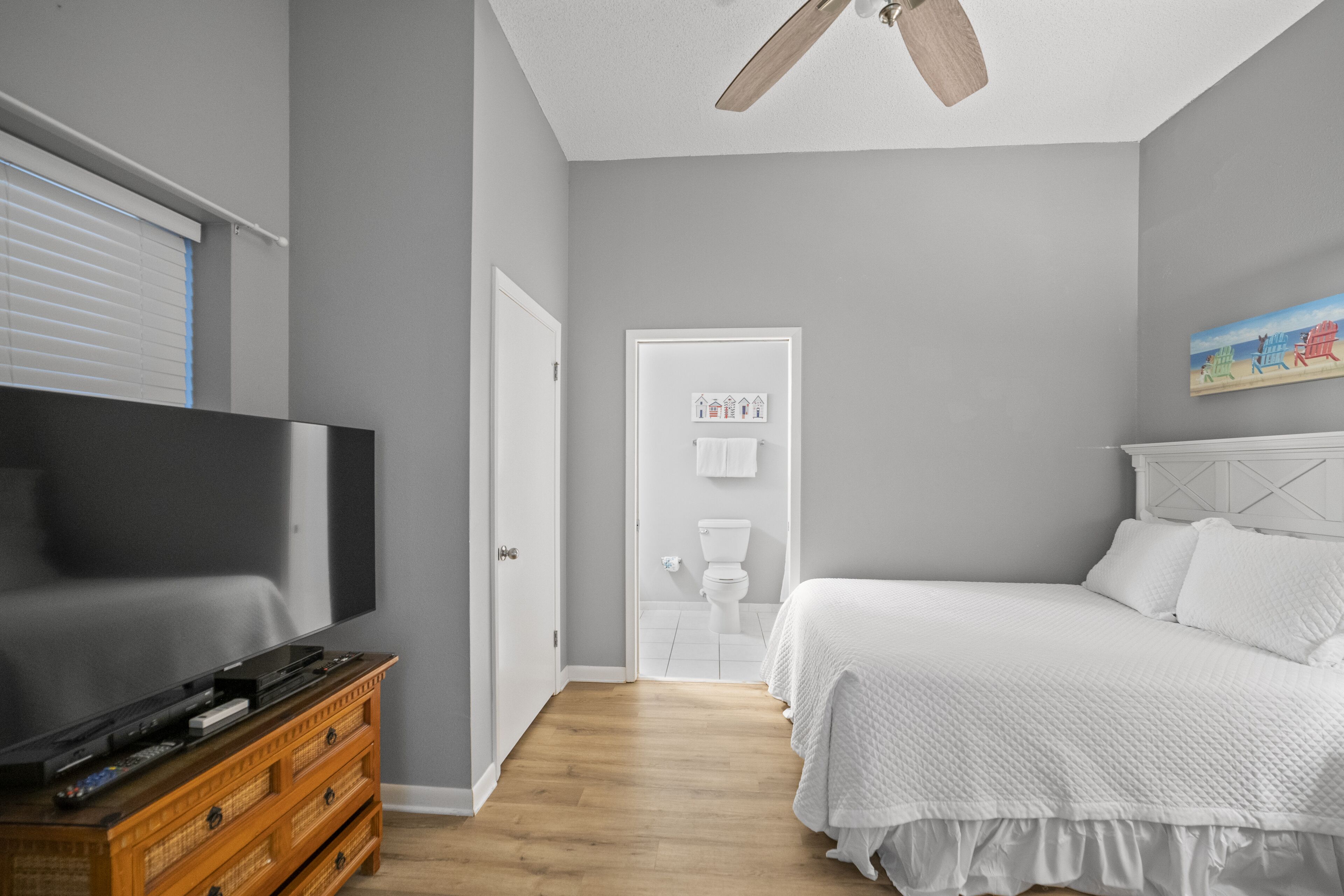 A bedroom with a TV on a wooden dresser, a bed with white bedding, and a ceiling fan.