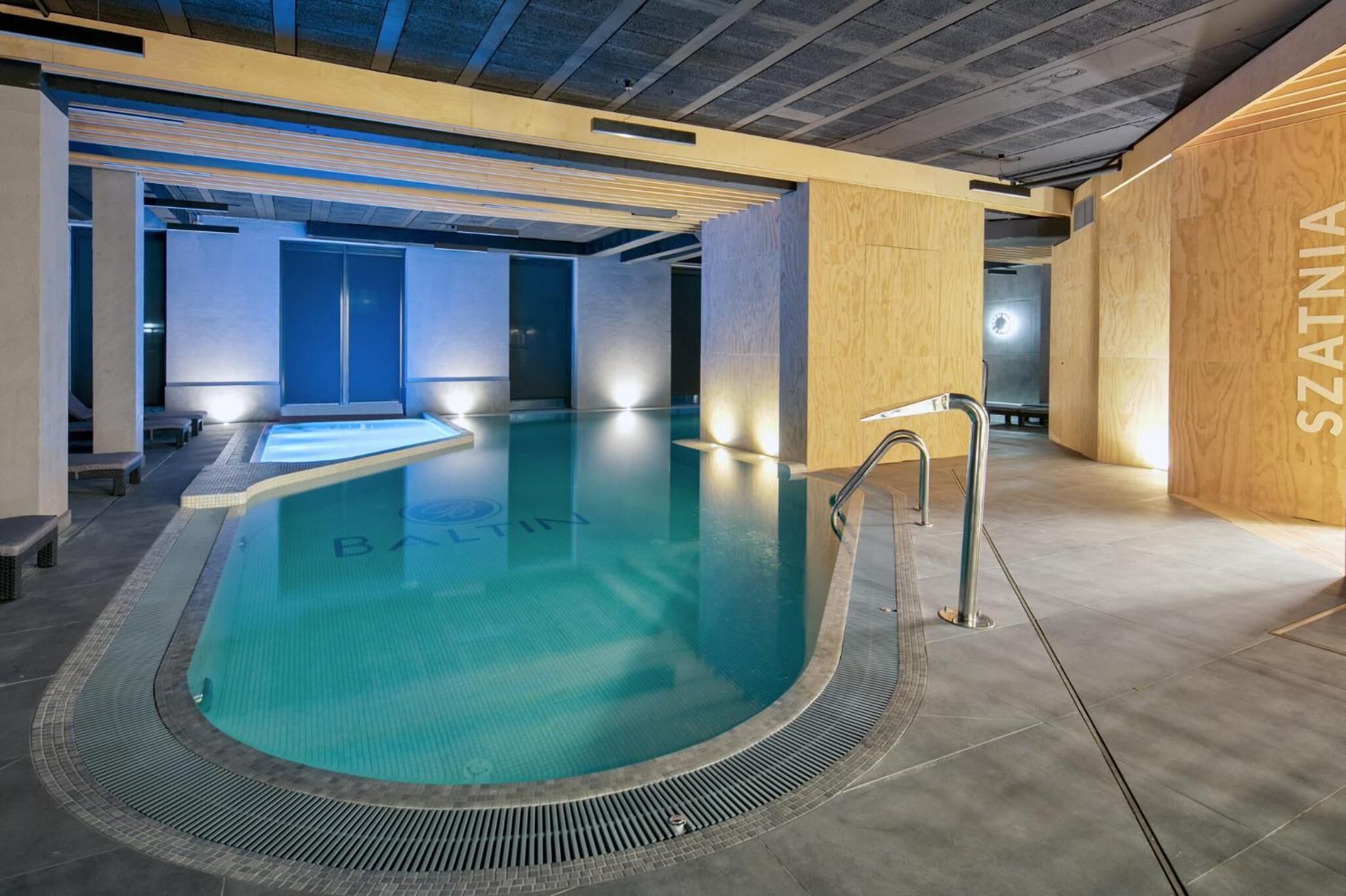 A modern indoor swimming pool with a clear blue water surface, surrounded by wooden walls and a tiled floor.