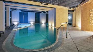A modern indoor swimming pool with a clear blue water surface, surrounded by wooden walls and a tiled floor.
