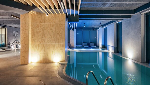 A modern indoor swimming pool with a wooden column, lounge chairs, and a clear ceiling with linear lighting. - Blu Apartments (Sarbinowo)